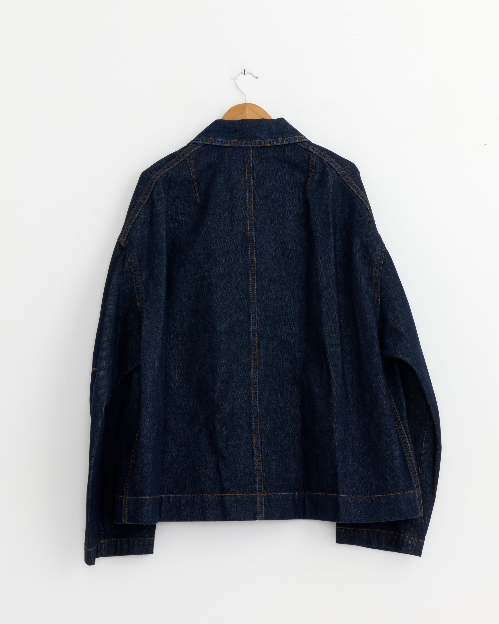 A Dries Van Noten Robinson Denim Coat in Indigo hangs on a wooden hanger against a plain white wall, displayed from the back.
