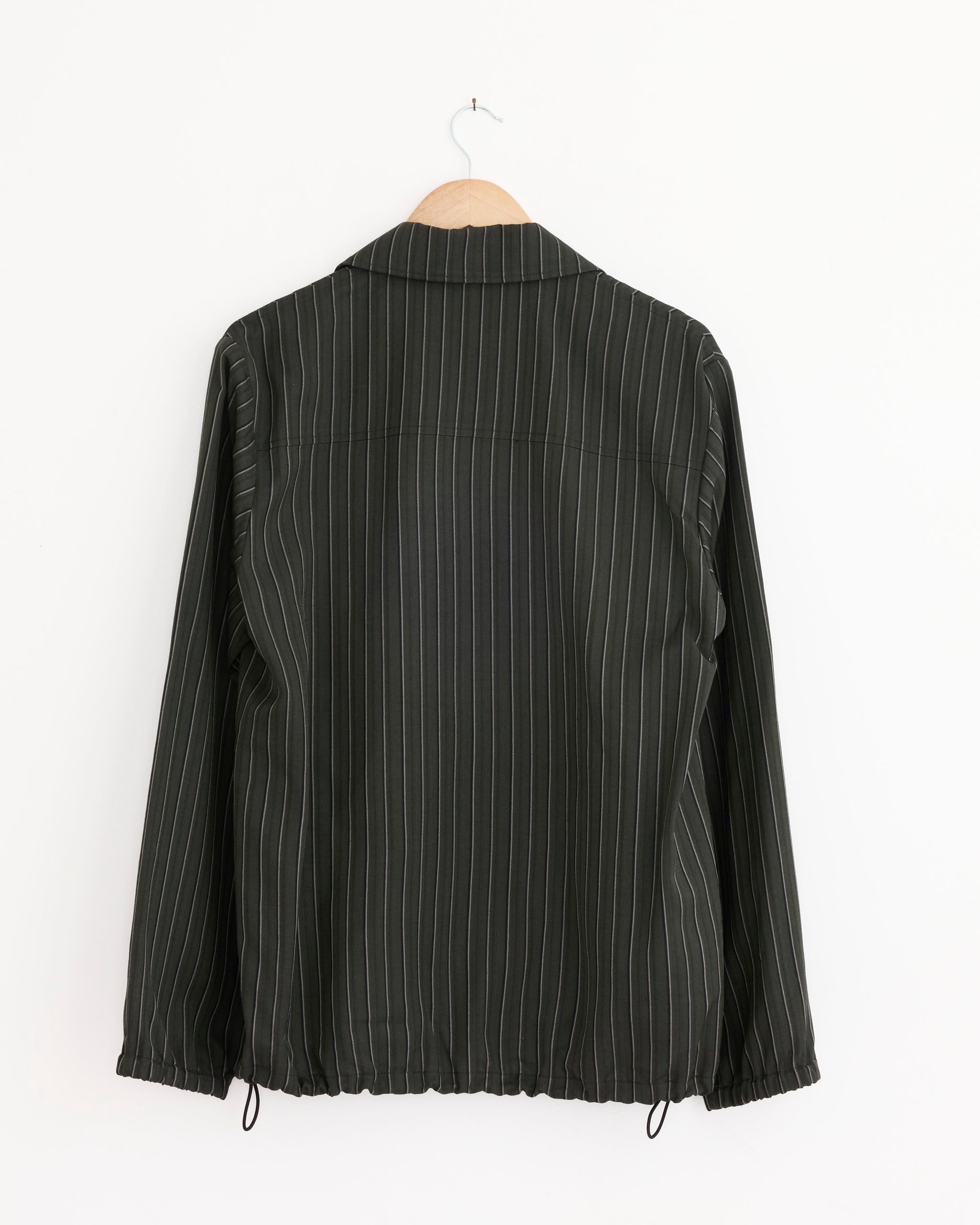 A Dries Van Noten Vorries Jacket in dark green hangs on a wooden hanger against a plain white wall, shown from the back.