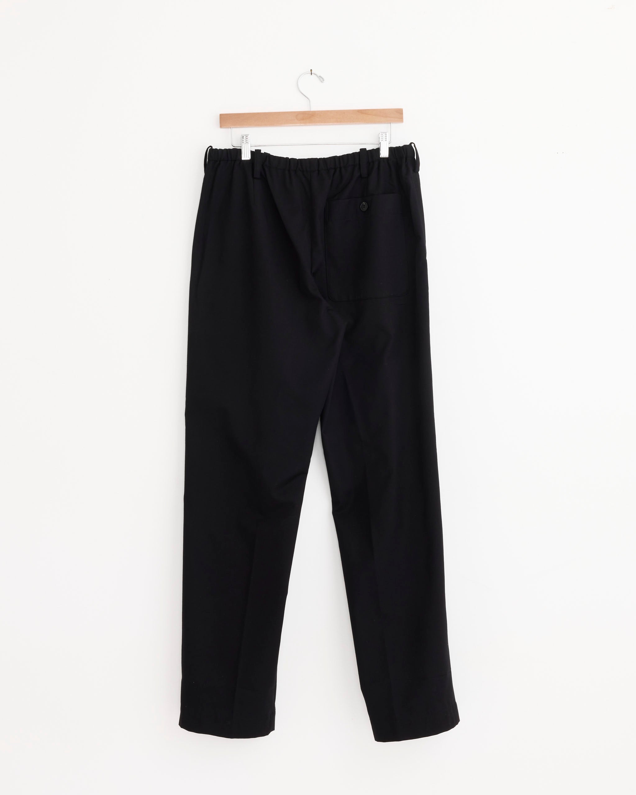 A pair of Dries Van Noten Penny Long Pant in Black, featuring an elastic waist and a buttoned back pocket, hangs on a wooden hanger against a white background.