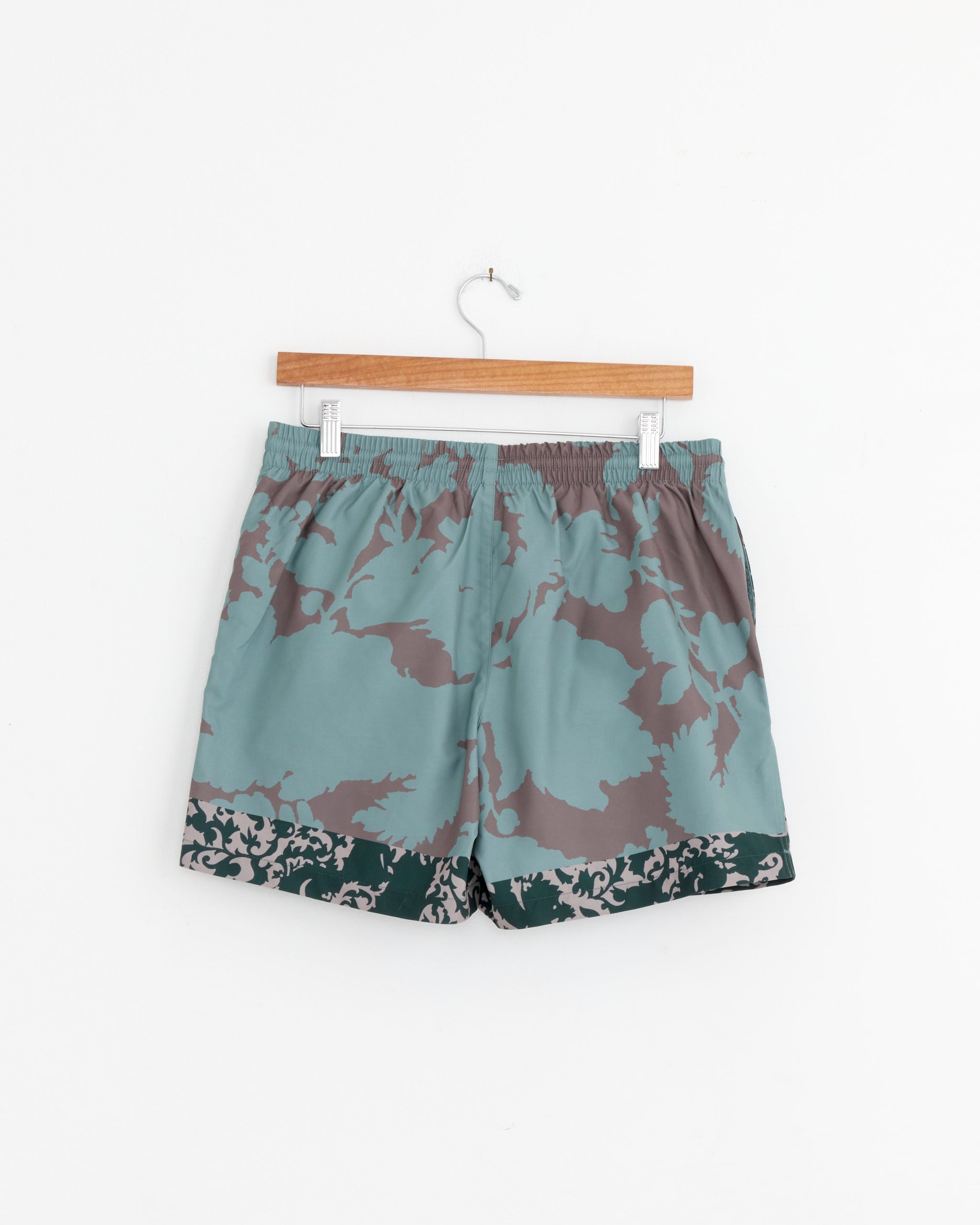 A pair of Dries Van Noten Swim Shorts in Petrol, featuring a teal, gray, and dark green camouflage pattern and elastic waistband, hangs on a wooden hanger against a white background.