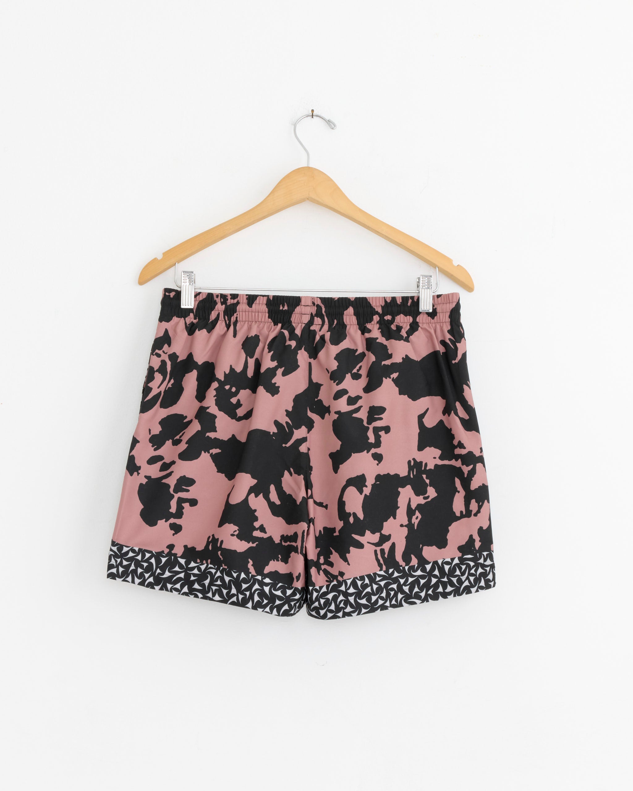 Dries Van Noten Swim Shorts in Old Rose hang on a wooden hanger against a white wall. These lightweight shorts feature an abstract print, elastic waistband, and geometric black-and-white border at the hem.