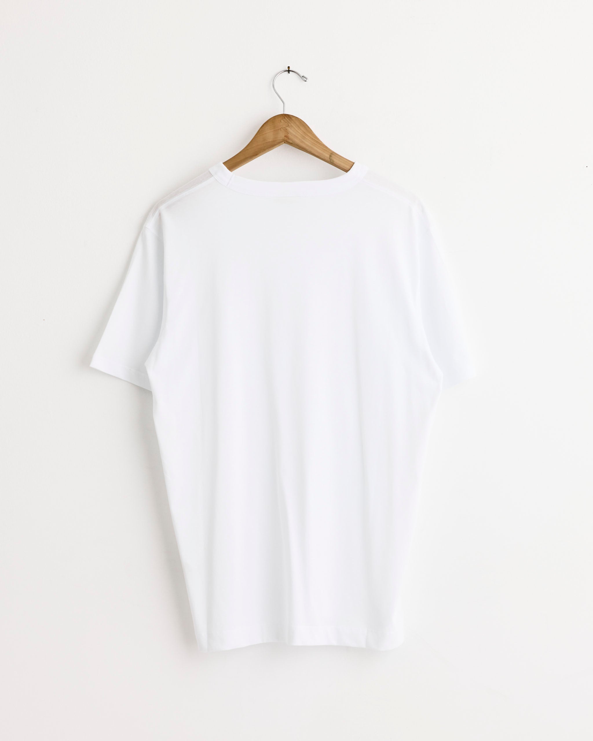 The Dries Van Noten Cotton T-Shirt in White, featuring an oversized fit and heavyweight cotton, hangs with its back facing forward on a wooden hanger against a white wall.