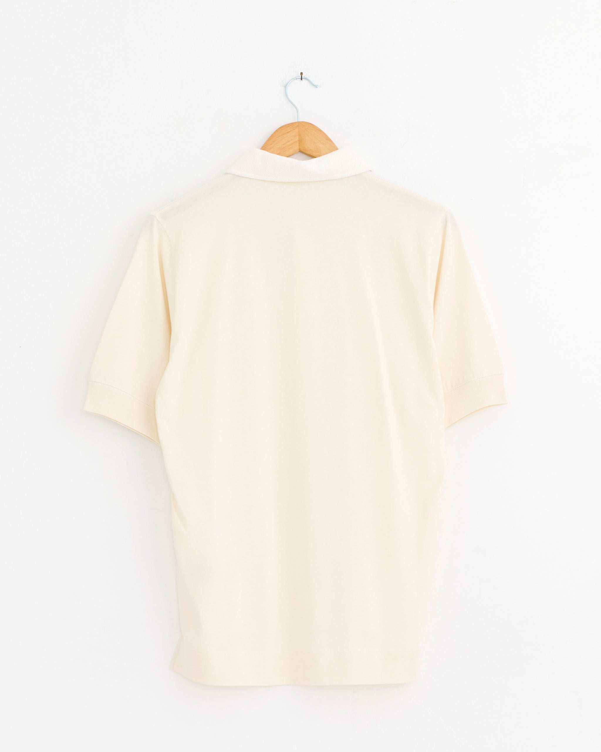 The Dries Van Noten Pointelle Polo in Off White, a short-sleeved jacquard knit shirt, hangs on a wooden hanger against a white wall, with its back facing the viewer.