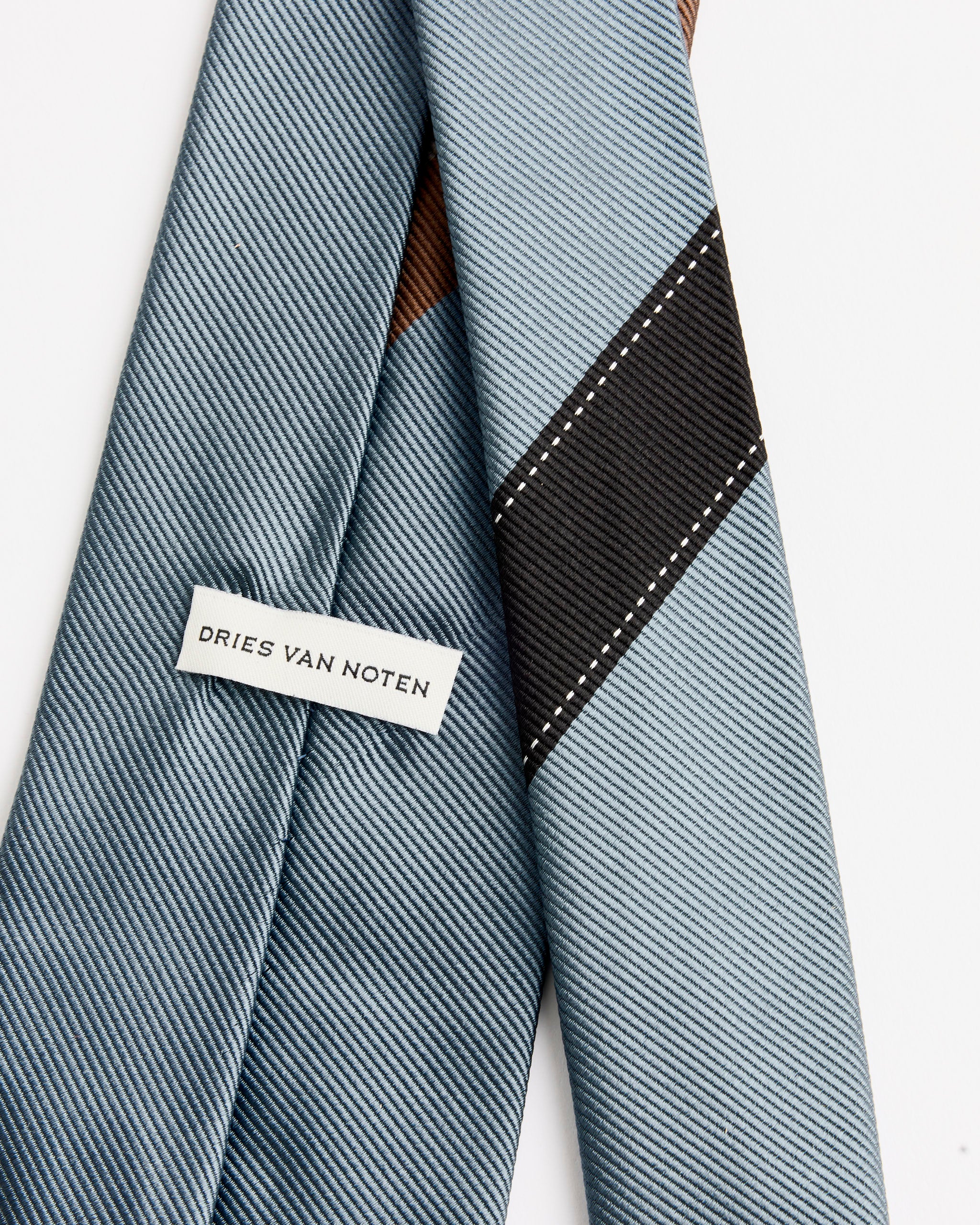 A close-up of the Dries Van Noten 3905 Tie in Raf shows light blue diagonal stripes with a black stripe and white stitching. A fabric label is sewn on the back, underscoring its luxurious 100% silk Italian craftsmanship.