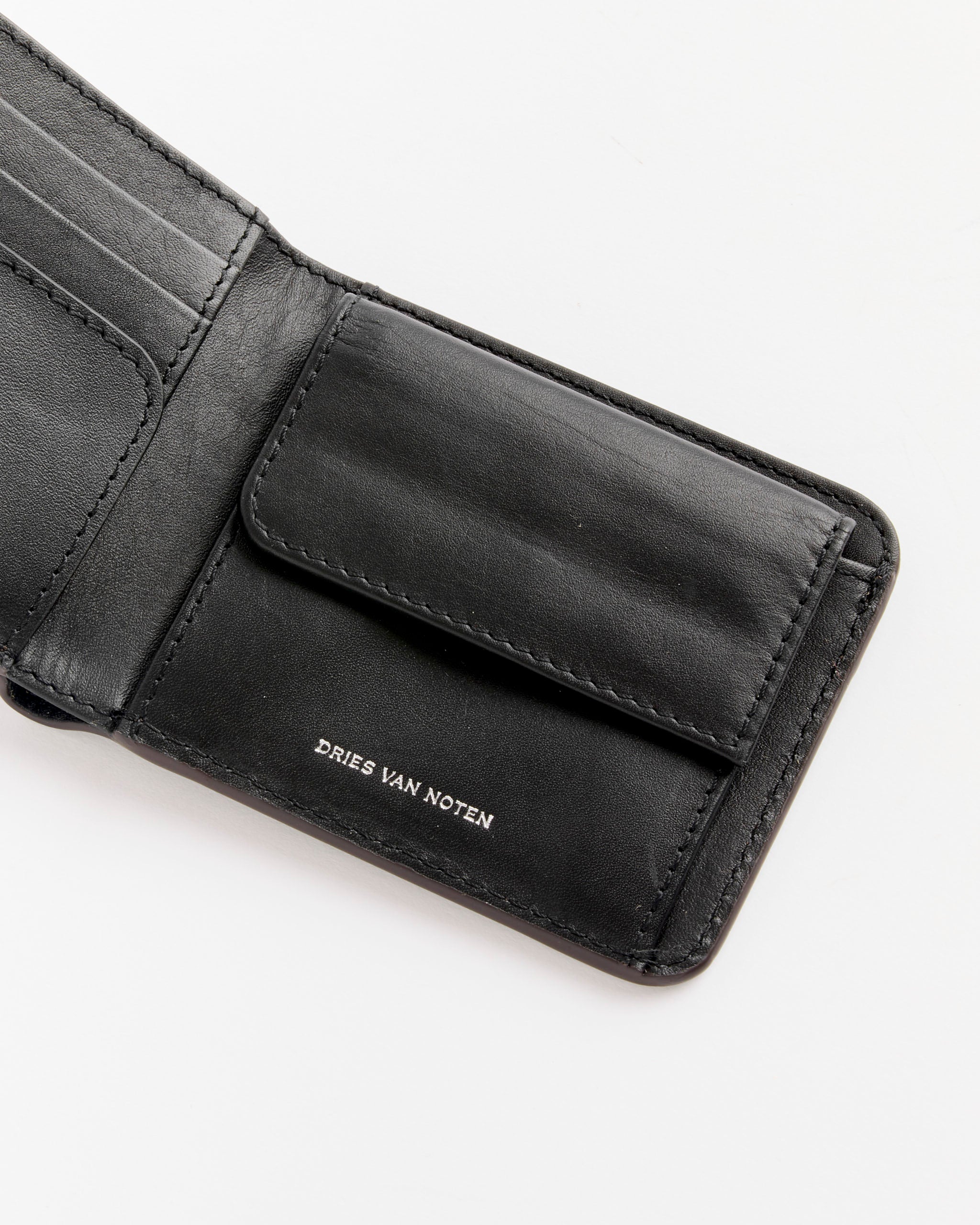 A dark grey bifold wallet by Dries Van Noten with multiple card slots and a coin pocket. "DRIES VAN NOTEN" is printed inside in small white letters. Shown on a white background.