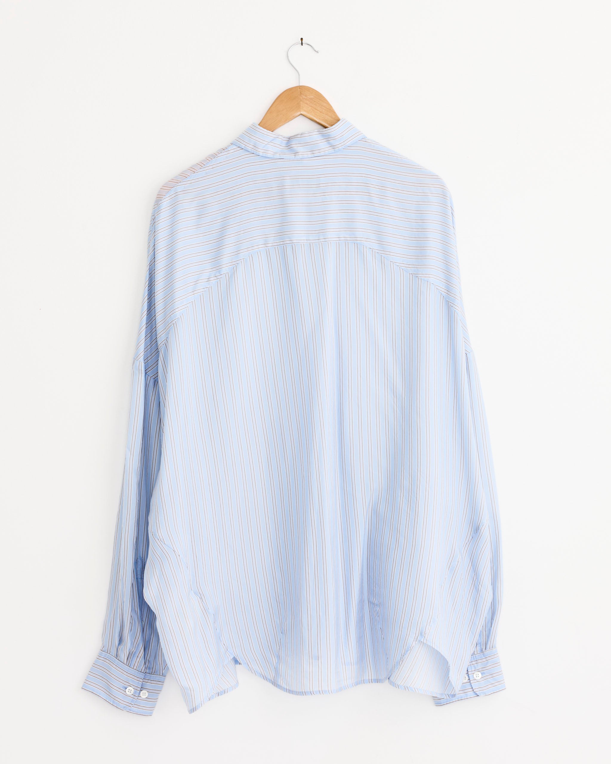 The Casia Striped Shirt in Light Blue by Dries Van Noten features an oversized fit with long sleeves and thin white and orange stripes, shown from the back on a wooden hanger against a plain white wall.