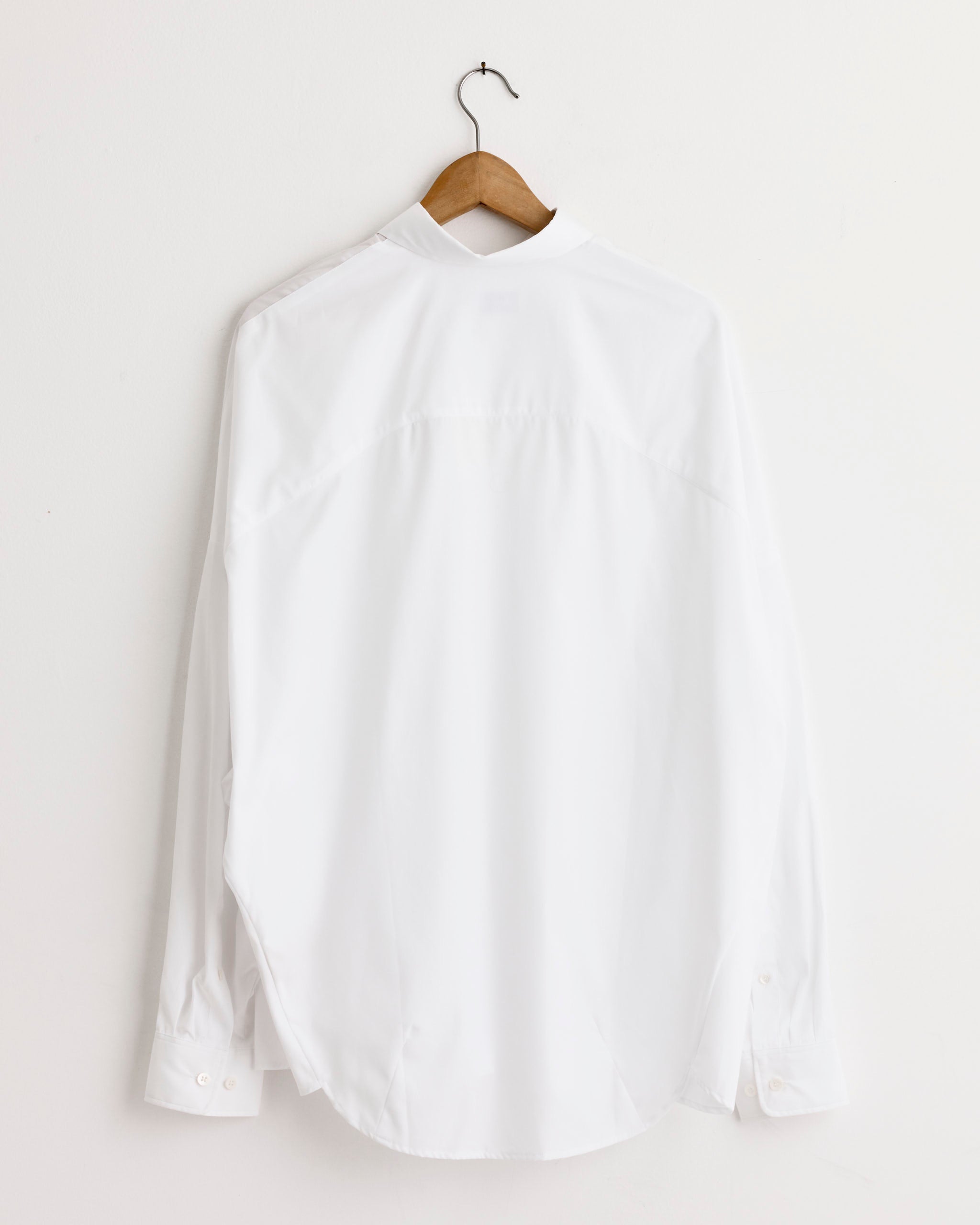 The Casia Shirt in White by Dries Van Noten—a crisp cotton oversized shirt with kimono sleeves—hangs on a wooden hanger, back facing forward, against a plain white wall.