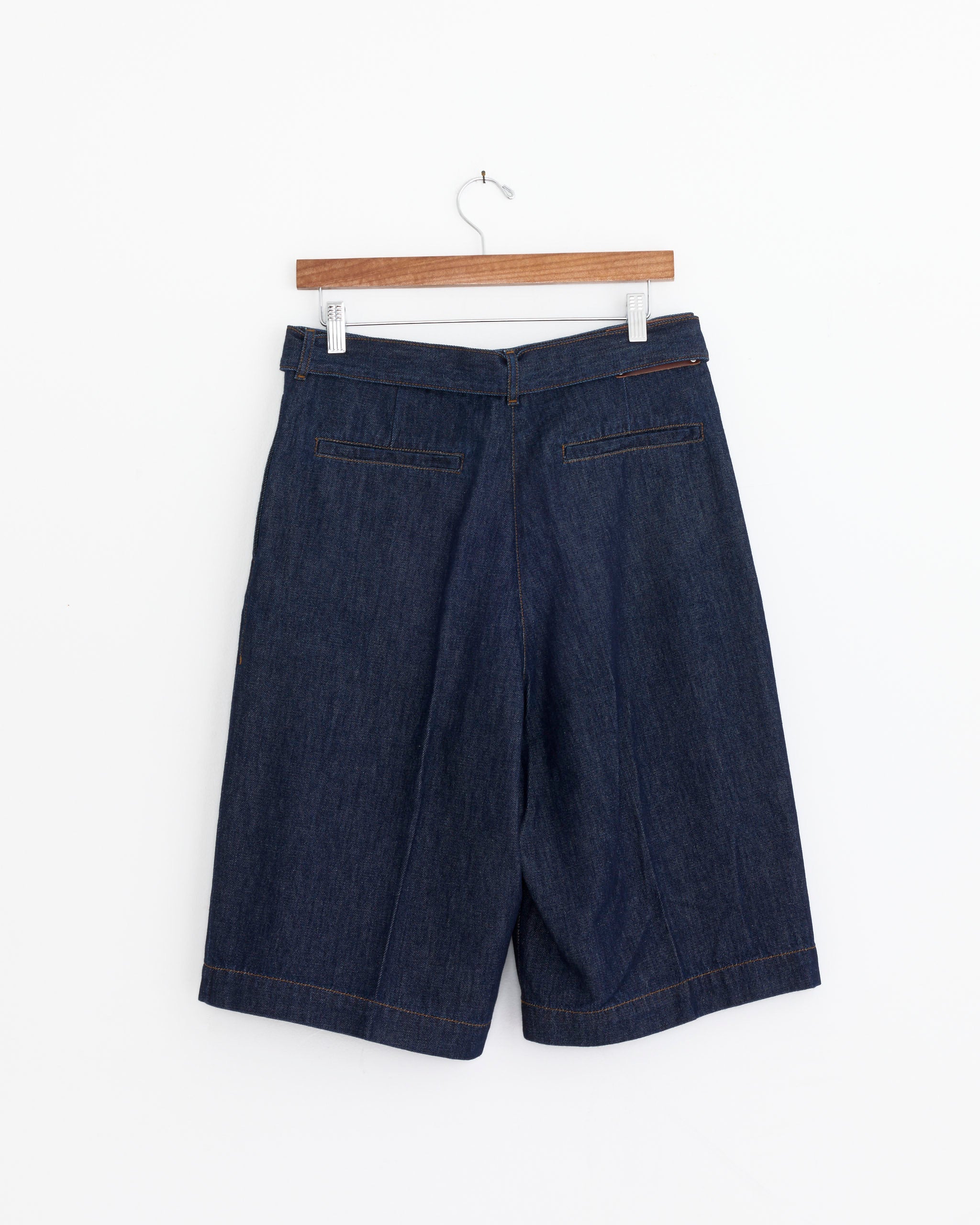 Dries Van Noten Belted Denim Shorts in Indigo hang on a wooden hanger against a white background, featuring two back welt pockets and visible stitching.
