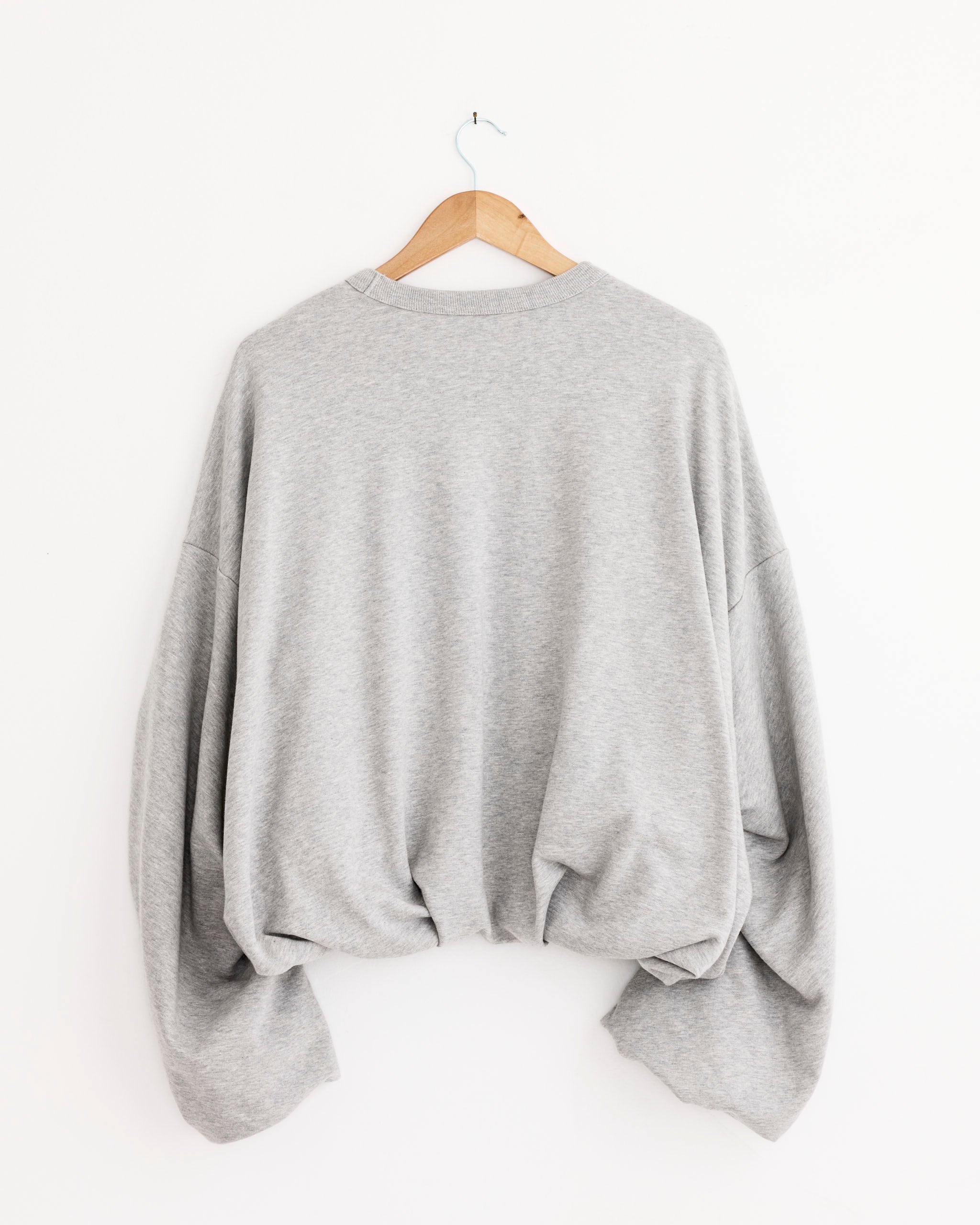 The Dries Van Noten Hannett Sweater in Grey Melange, featuring long sleeves and an oversized fit, hangs on a wooden hanger against a white wall, showcasing its soft fabric and relaxed, draped silhouette.
