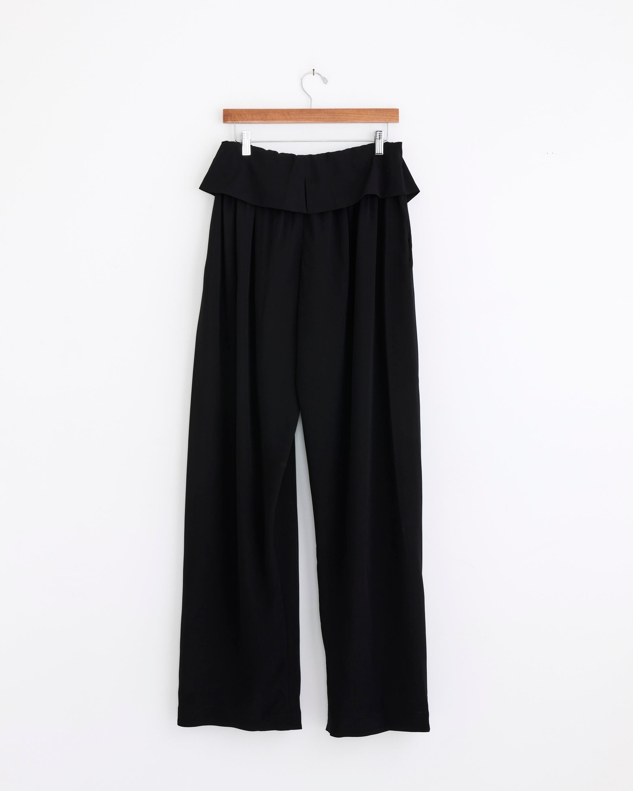 The Dries Van Noten Draped Trousers in Black feature a high waist, wide legs, and a ruffled waistband, displayed on a wooden hanger against a plain white background.