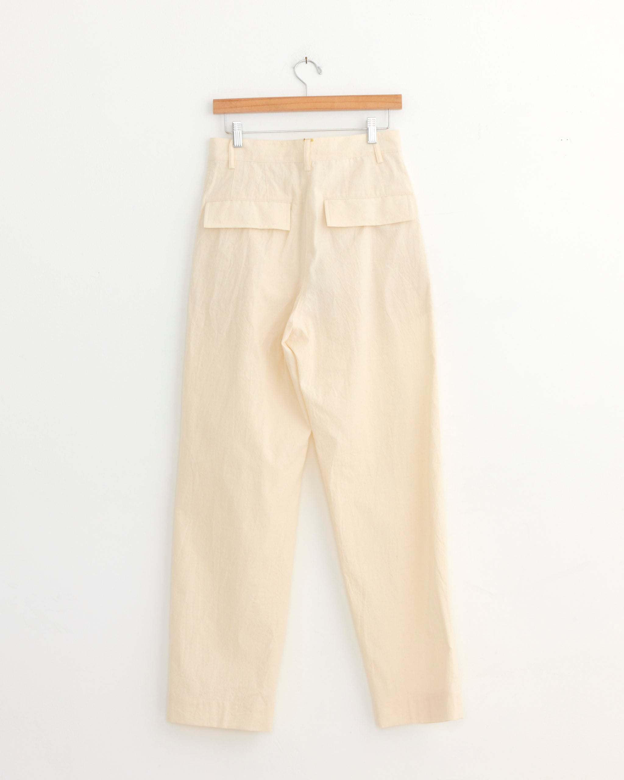 The Dries Van Noten Wide-Leg Pant in Ecru features a high waist, straight wide legs, and two back flap pockets, displayed on a wooden hanger against a plain white wall.