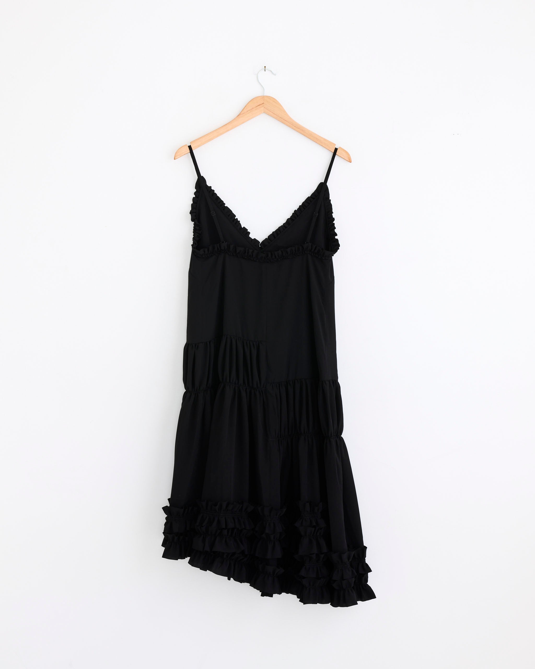 The Dries Van Noten Ruffled Midi Dress in Black, featuring spaghetti straps and a ruffled hem, hangs on a wooden hanger against a plain white wall, capturing effortless elegance.