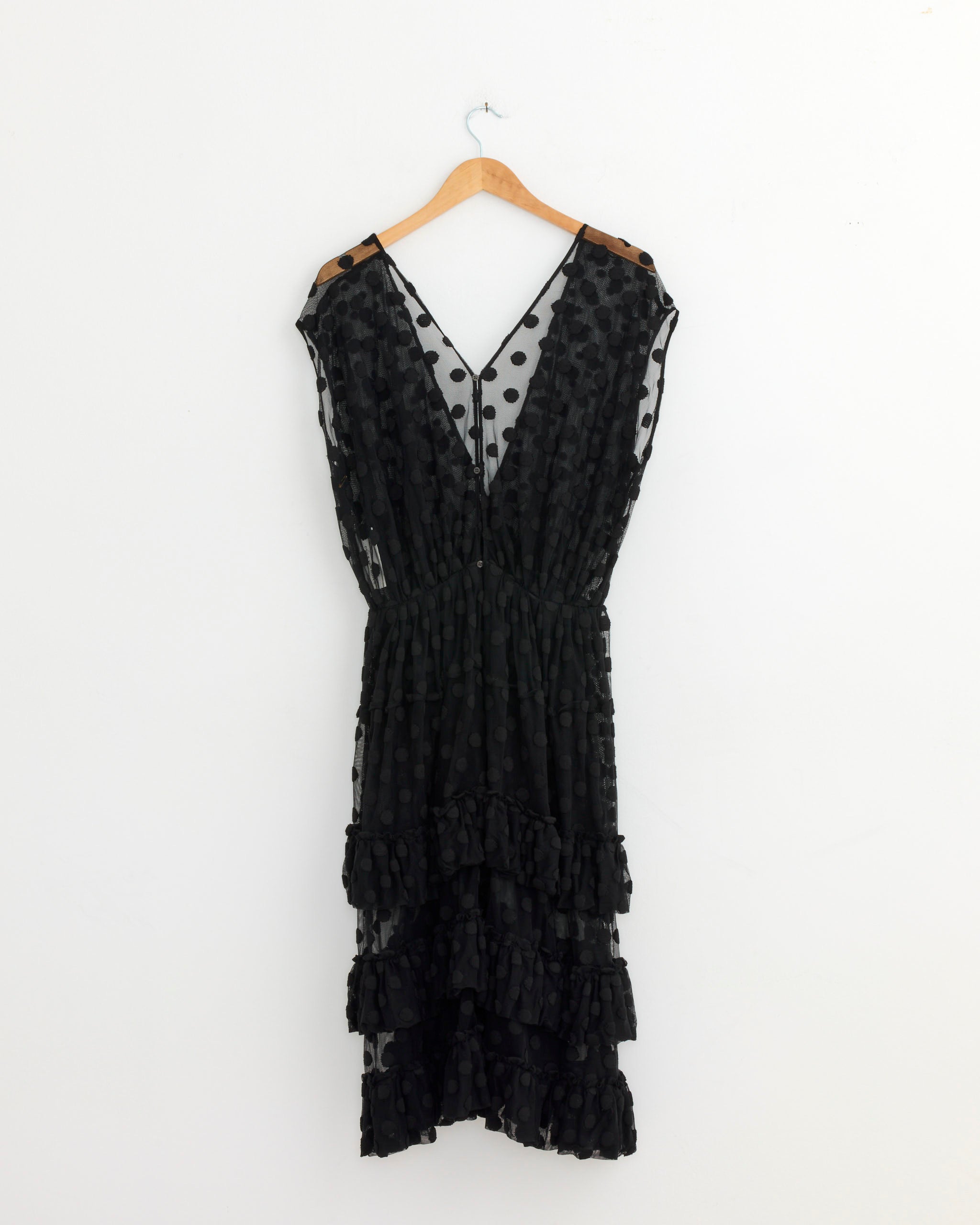The Mesh Ruffle Dress in Black by Dries Van Noten, featuring a sleeveless design, polka dots, sheer mesh fabric, deep V-neckline, and layered ruffles on the skirt, hangs on a wooden hanger against a white wall.