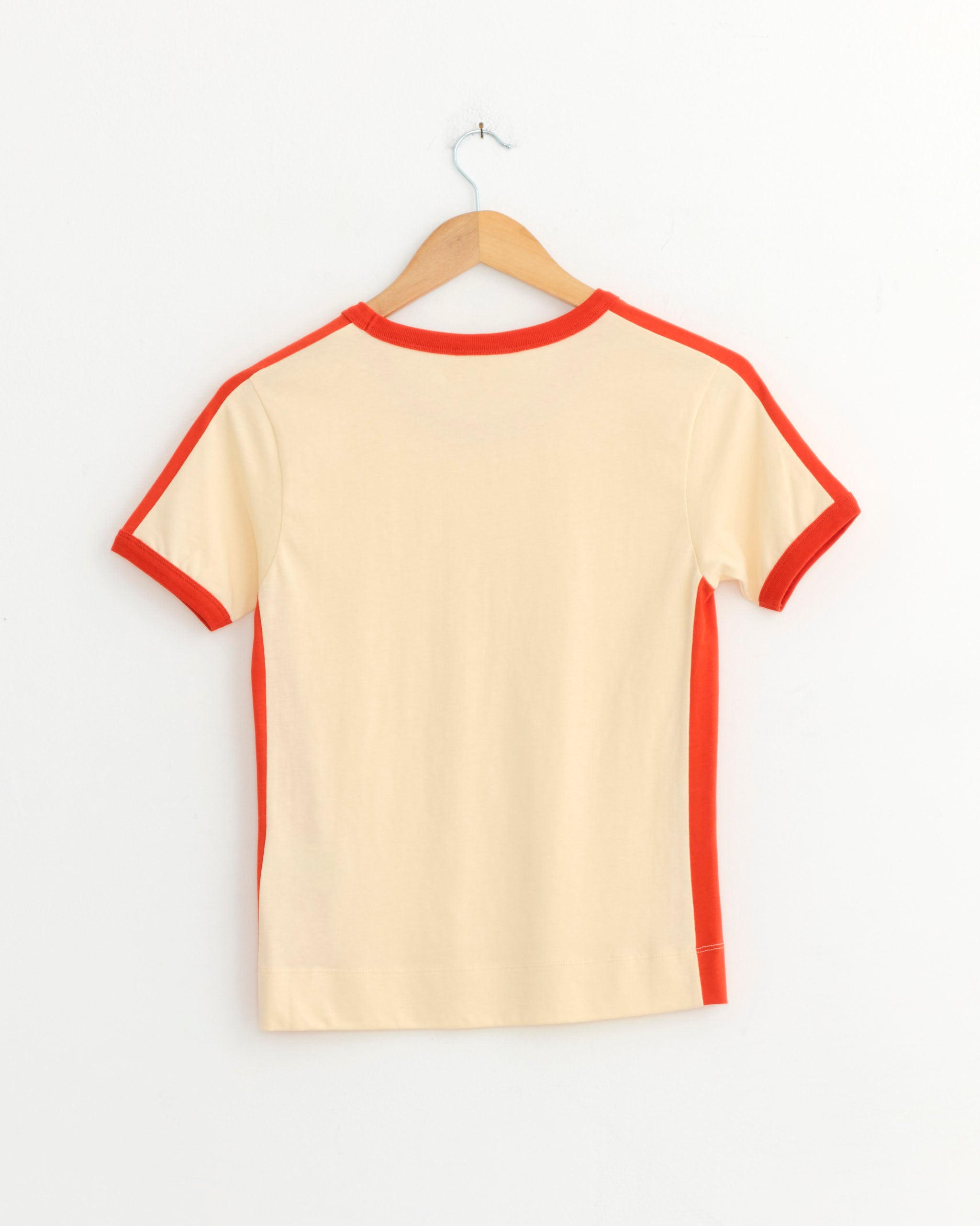 The Dries Van Noten Contrast T-Shirt in Ivory, crafted from soft cotton jersey with red trim on the collar, cuffs, and side seams, hangs on a wooden hanger against a white wall.