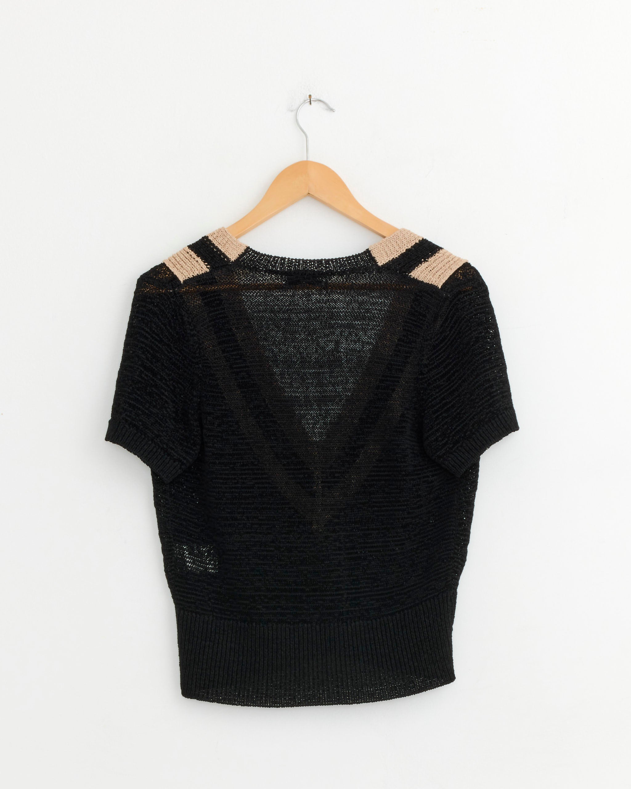 The Striped Knitted Sweater in Black by Dries Van Noten, featuring a deep V-shaped sheer pattern and beige shoulder accents, hangs on a wooden hanger against a white wall.