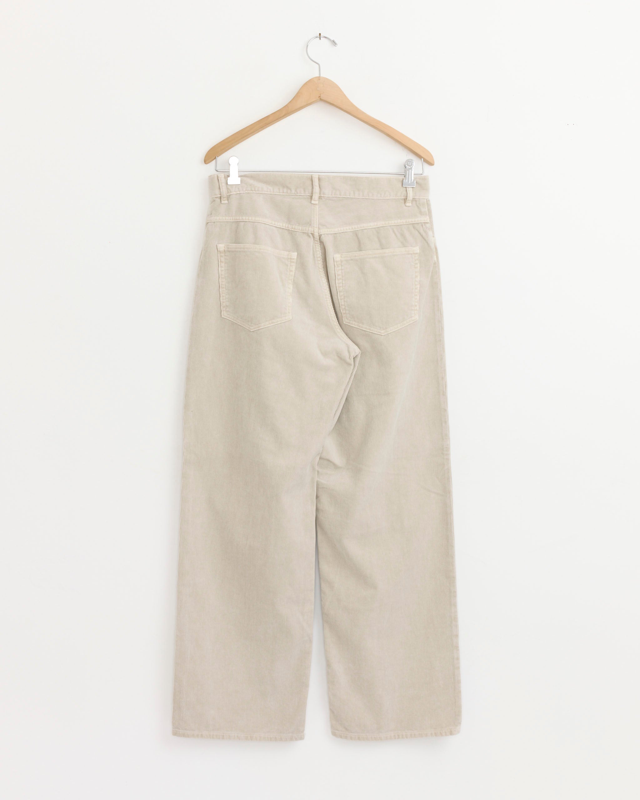 The Garment Dyed Corduroy 5 Pocket Pant in White Khaki by Auralee is shown from the back on a wooden hanger against a white background, featuring two rear pockets and wide, straight legs in a relaxed fit.