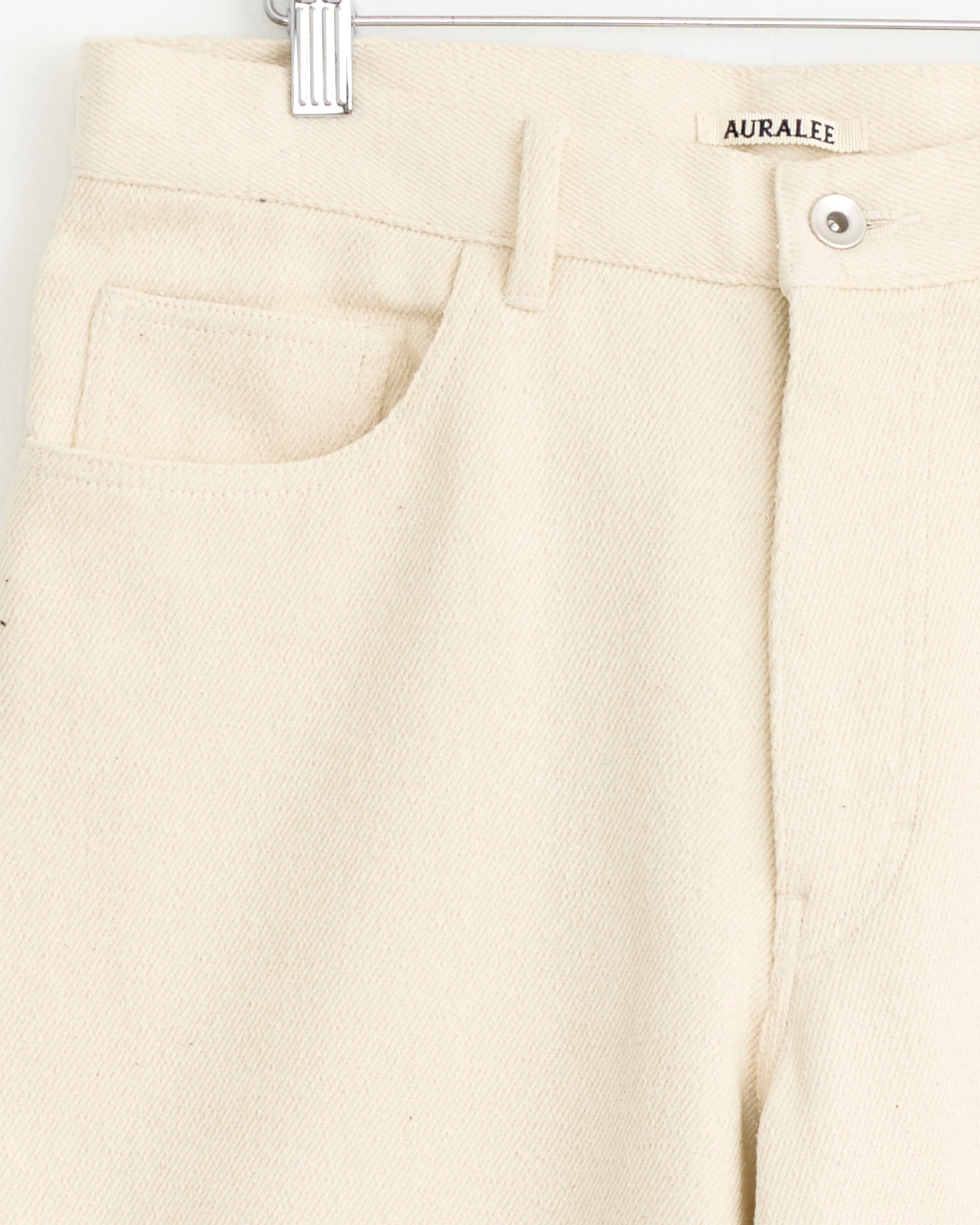 Close-up of Auralee Silk Nep Denim 5 Pocket Short in Ivory, made in Japan, featuring front pocket, belt loop, button, visible brand label on waistband. Cream-colored wide fit denim hangs on a hanger against a white background.