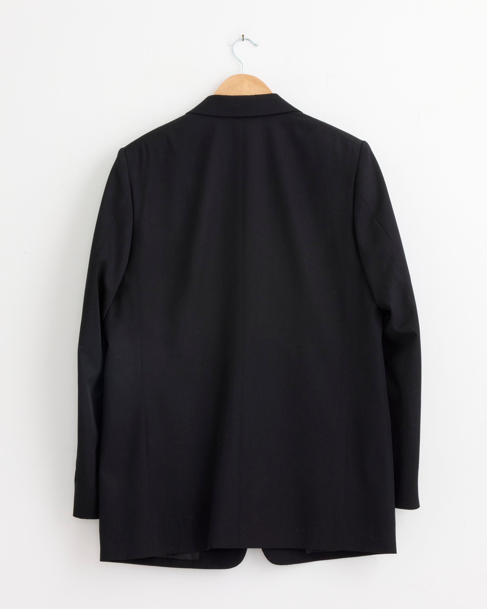 The Auralee Light Wool Max Gabardine Jacket in Top Black, made from Super 140’s Wool, hangs on a wooden hanger against a plain white wall, shown from the back.