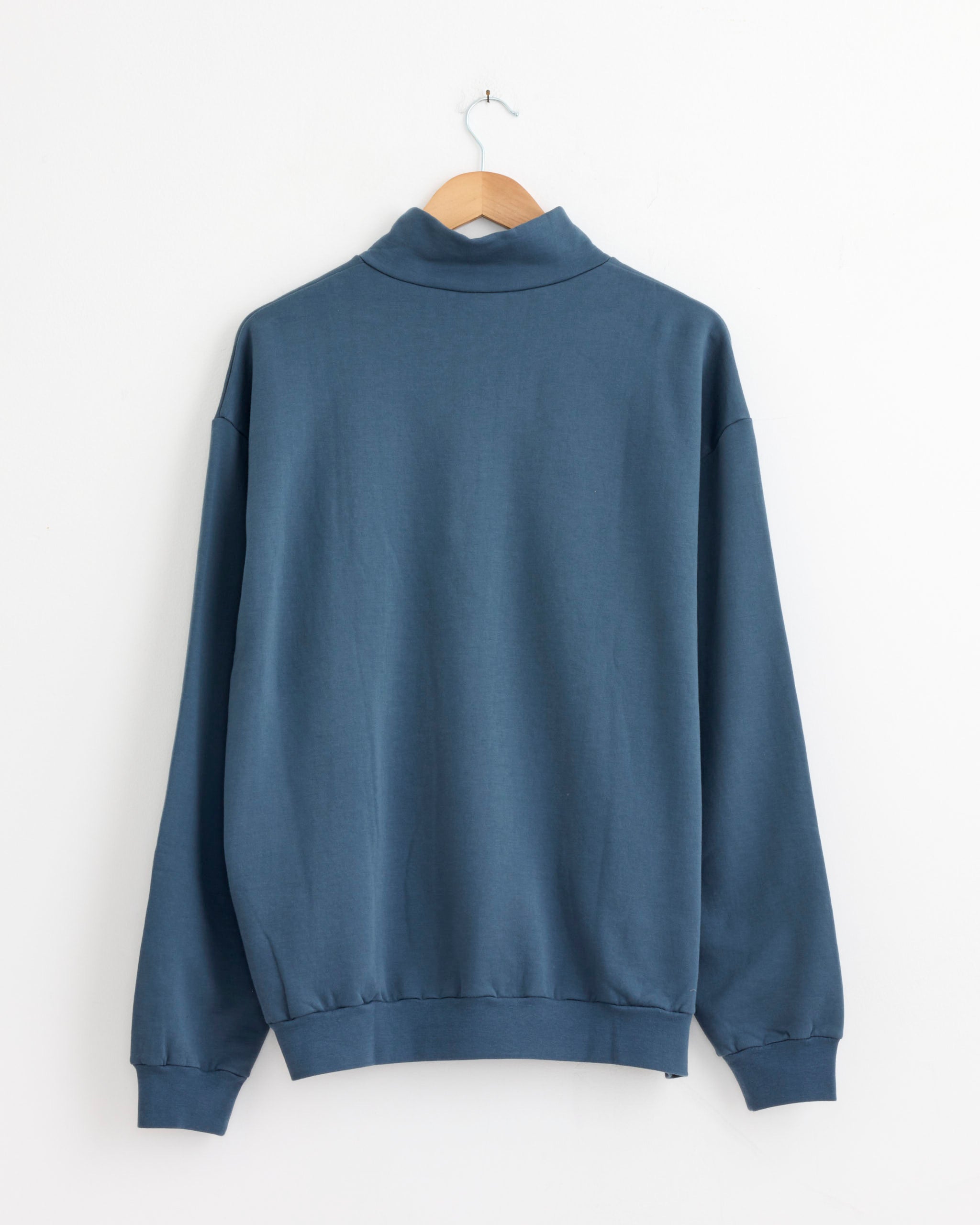 The Auralee Elastic High Gauge Sweat Zip Pullover in Dusty Blue, made from extra-long staple cotton for premium comfort and durability, is displayed on a wooden hanger against a white wall, shown from the back. Made in Japan.