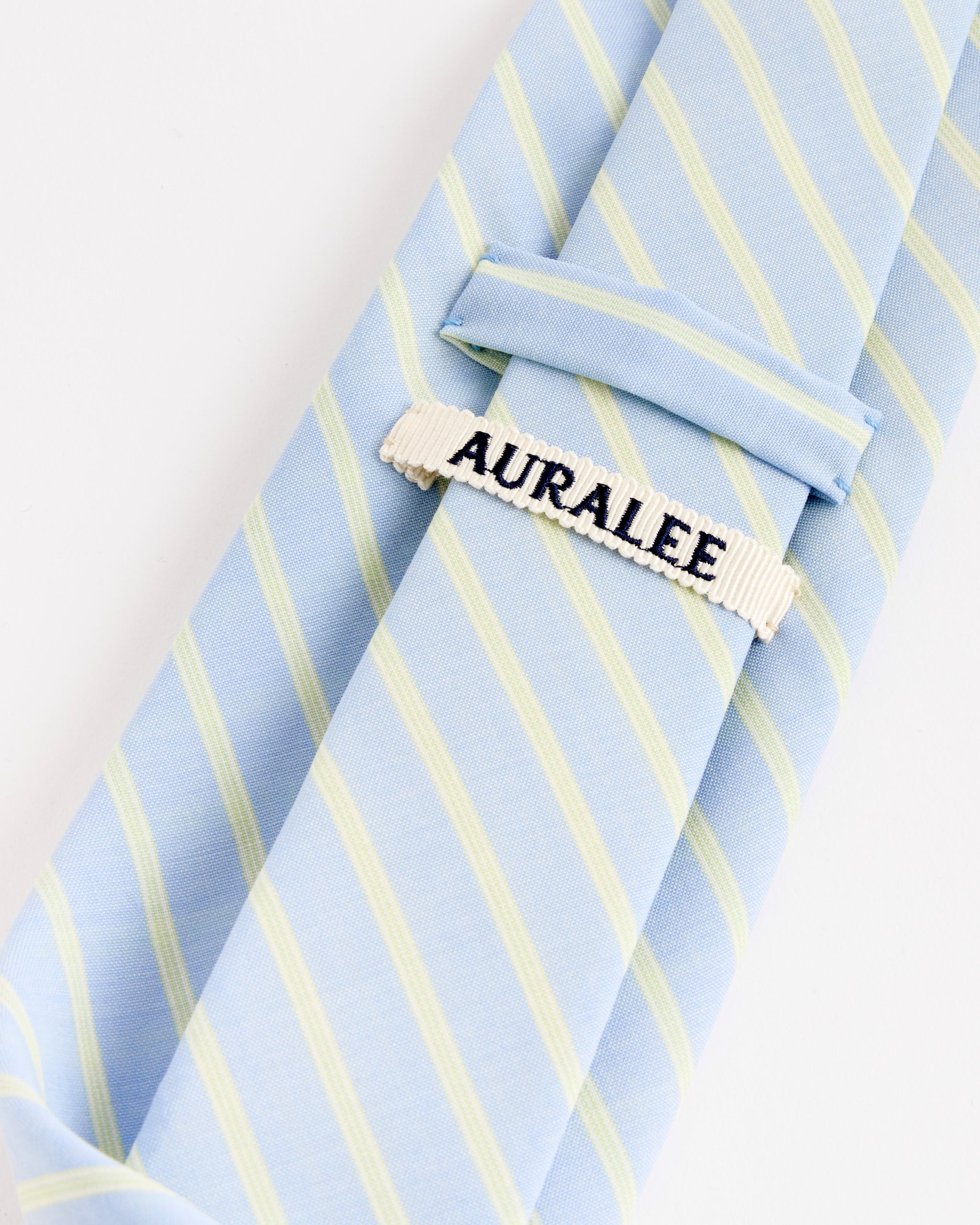 The Super Fine Wool Stripe Tie in Sax Blue by Auralee features pale yellow stripes and a white tie bar with "AURALEE" embroidered in black. Crafted from luxurious Super 180’s Wool for an elegant finish.