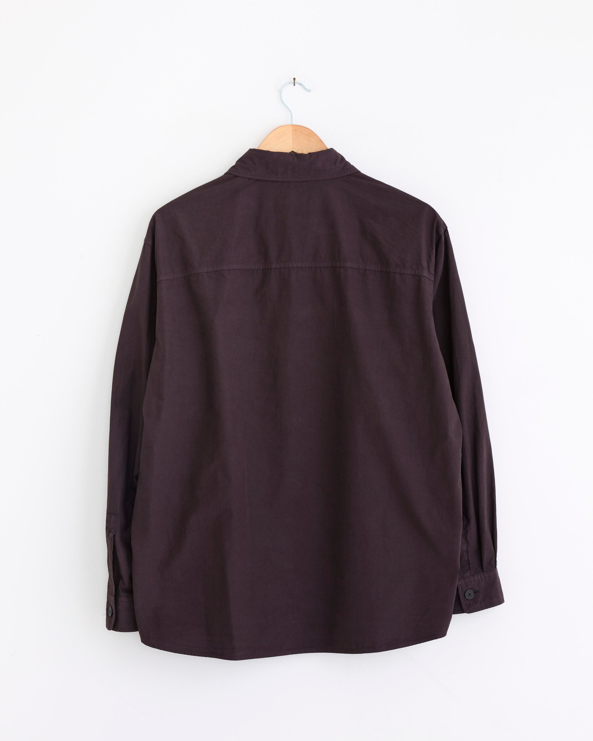 An Auralee Garment Dyed Finx Shuttle Shirt in Dark Brown, crafted in Japan, is displayed from the back on a wooden hanger against a plain white wall.