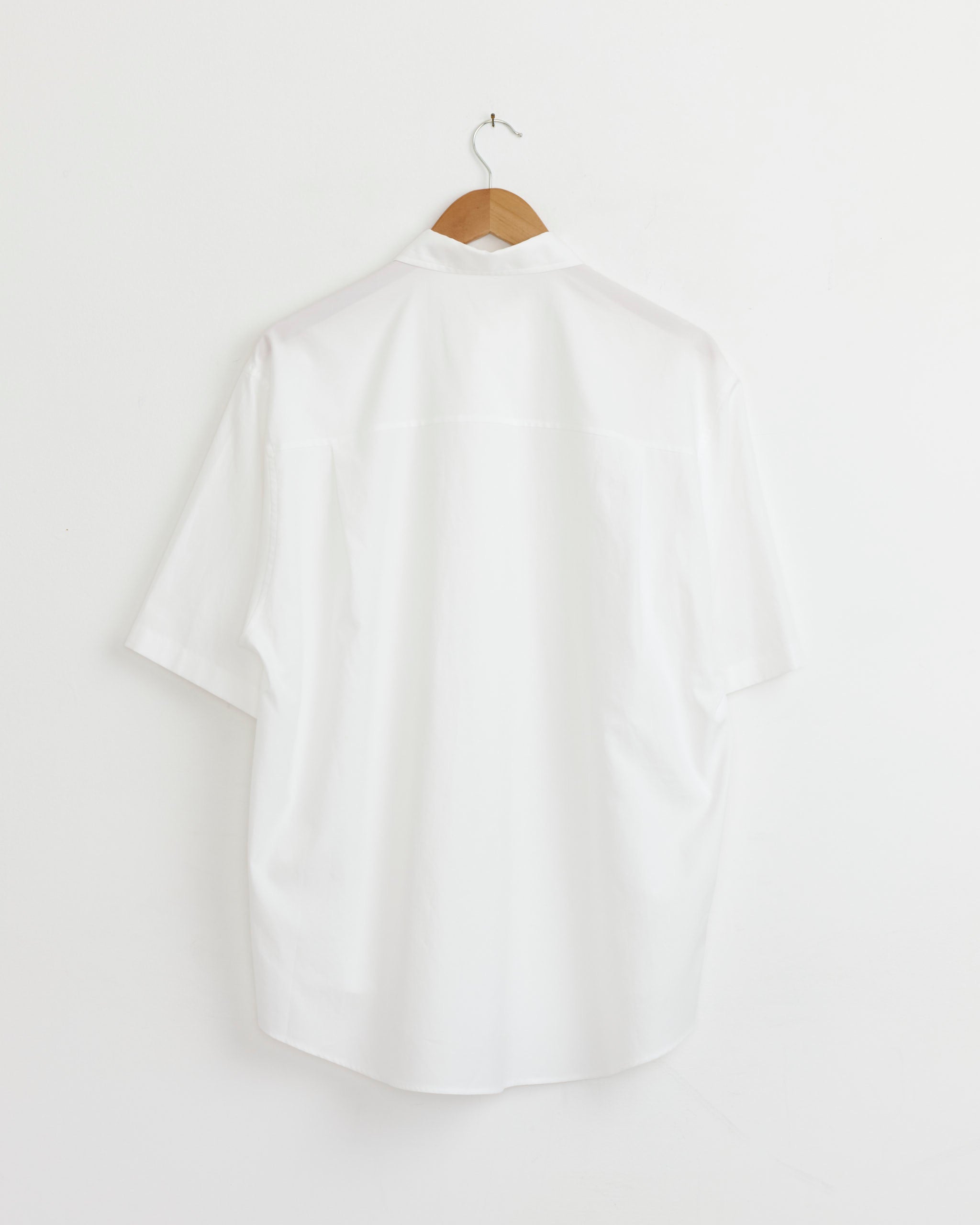 The Auralee Washed Finx Twill Big Shirt in White, shown from the back, hangs on a wooden hanger against a white wall.