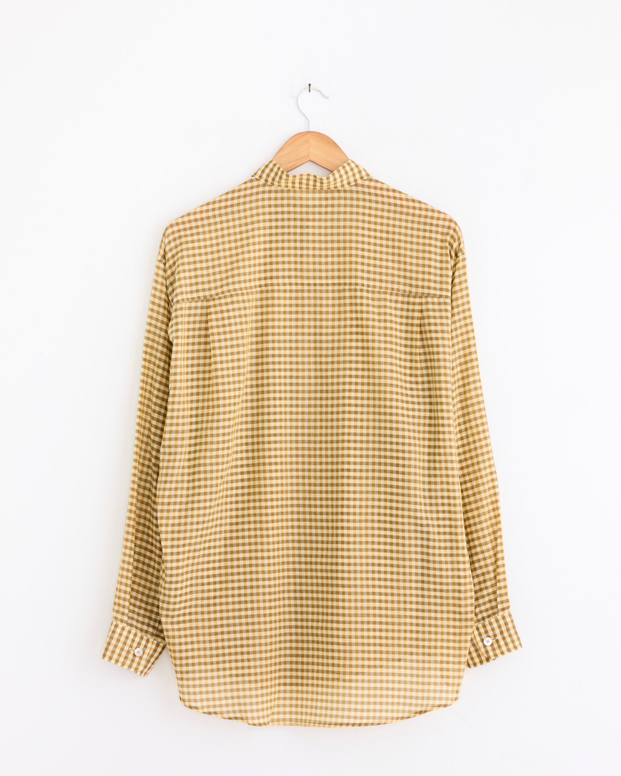 The Auralee Airy Light Cotton Check Big Shirt in Mustard Gingham, an oversized long-sleeve cotton piece, is displayed from the back on a wooden hanger against a plain white wall.