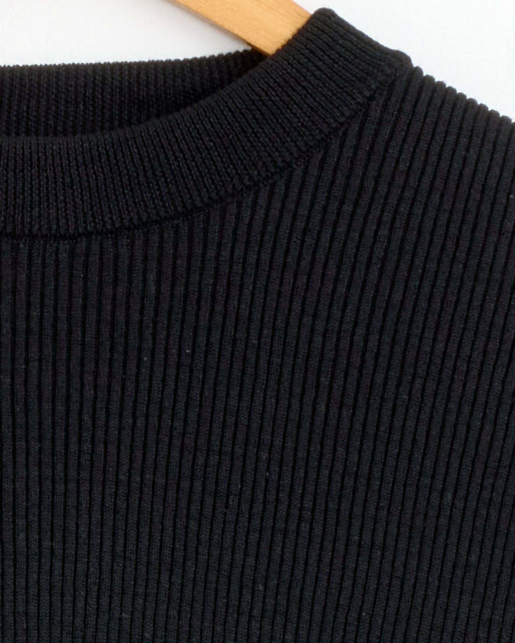 Close-up of Arpenteur's Standard Knit in Black, a ribbed sweater crafted from a linen-cotton blend, shown on a wooden hanger with focus on the neckline and vertical textured stripes.
