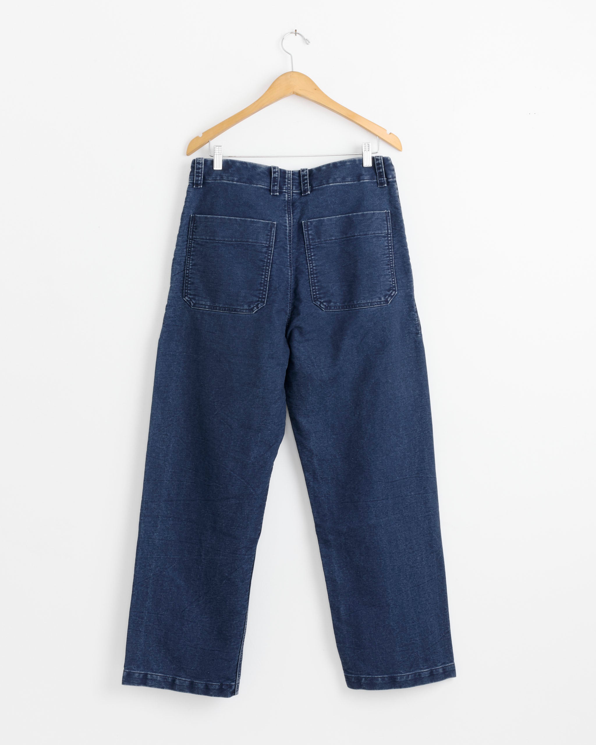 A pair of Arpenteur Lux Pant in Indigo, made in France from cotton, hangs on a wooden hanger against a white background, displayed from the back to show large back pockets.