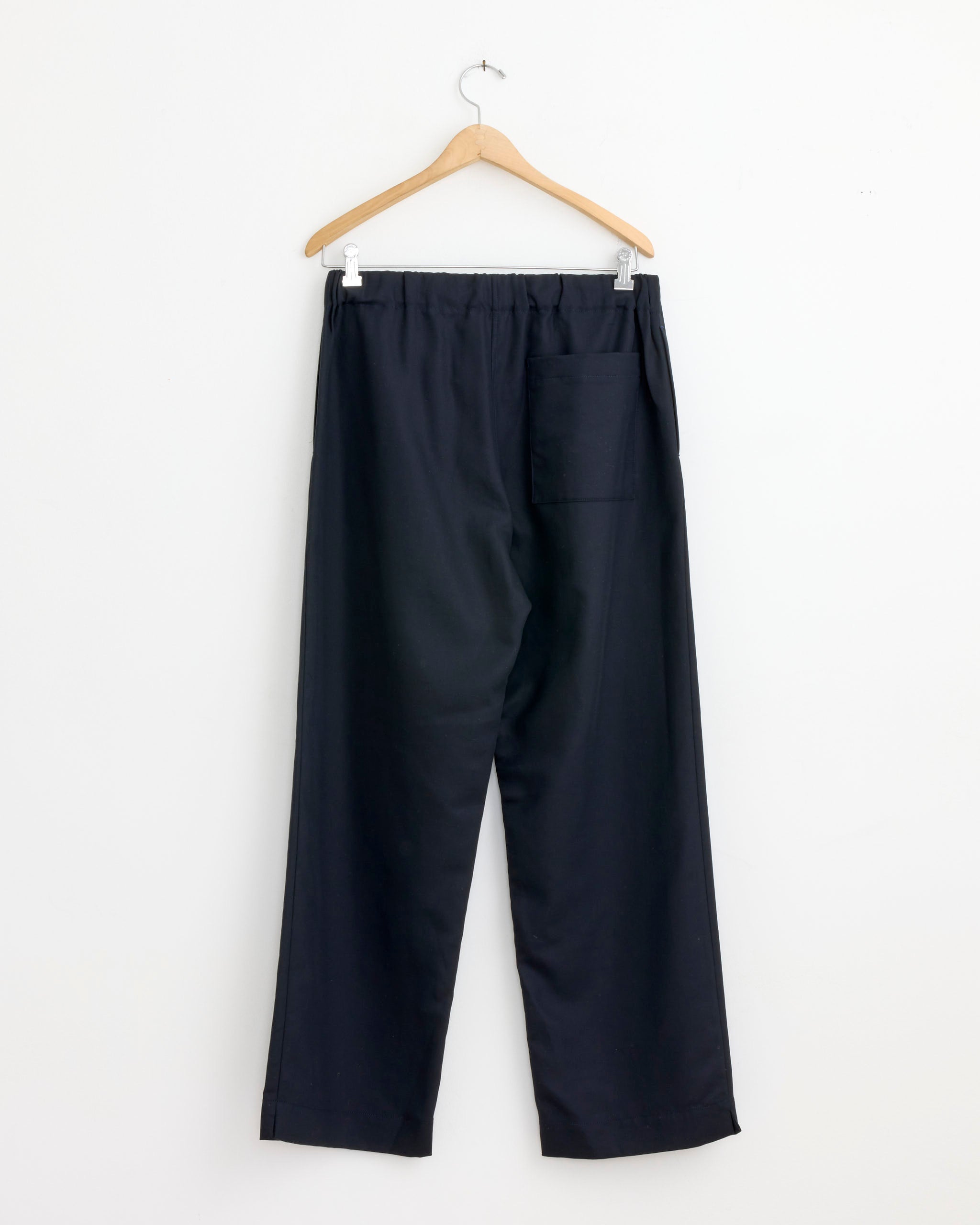 The Arpenteur Repos Pant in Midnight, made in France, has a loose fit, elastic waistband, and single back pocket, shown on a wooden hanger against a white wall.