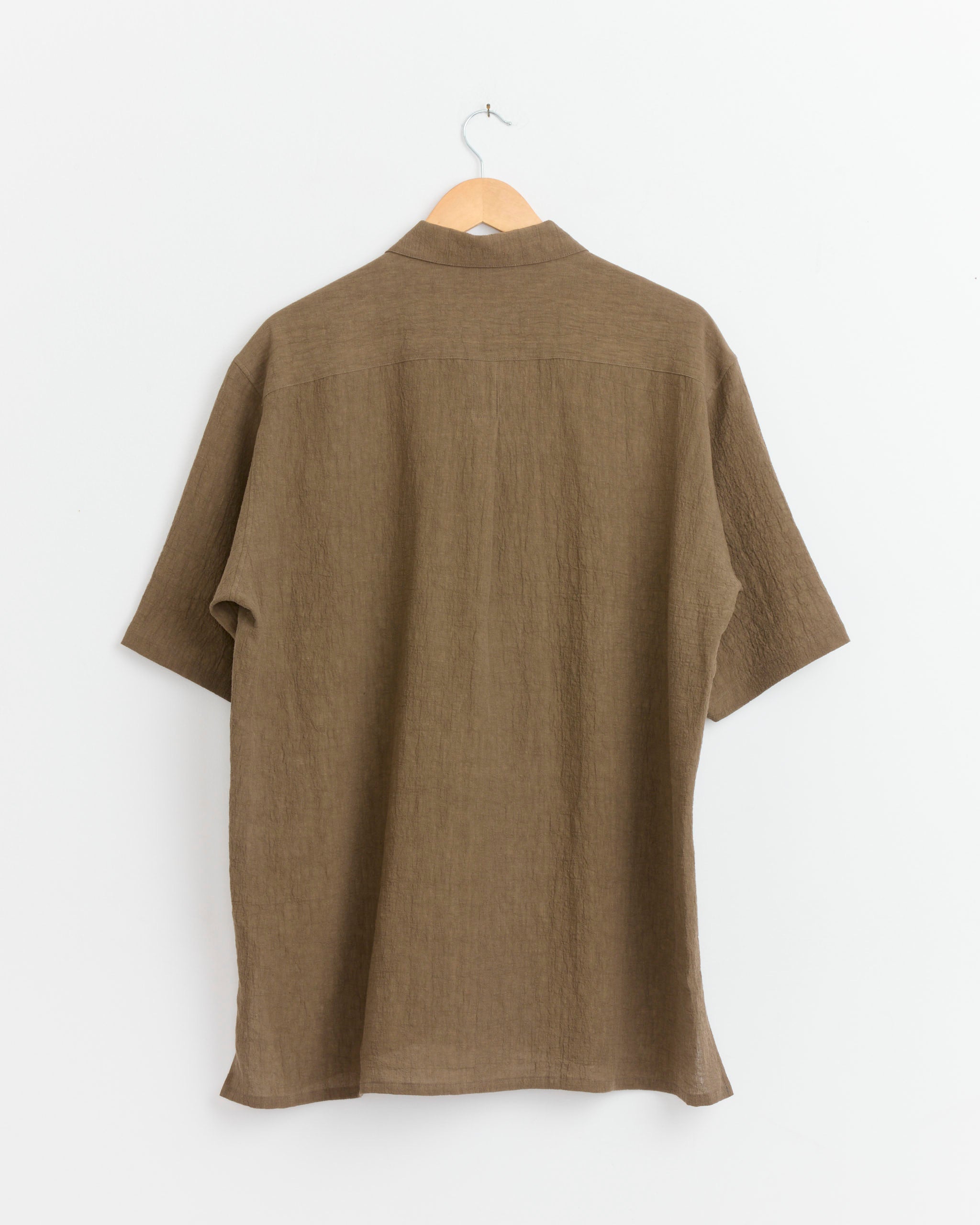 The Cale Water Twisted Linen Shirt in Brown, made in Japan, hangs on a wooden hanger against a plain white wall, displayed from the back.
