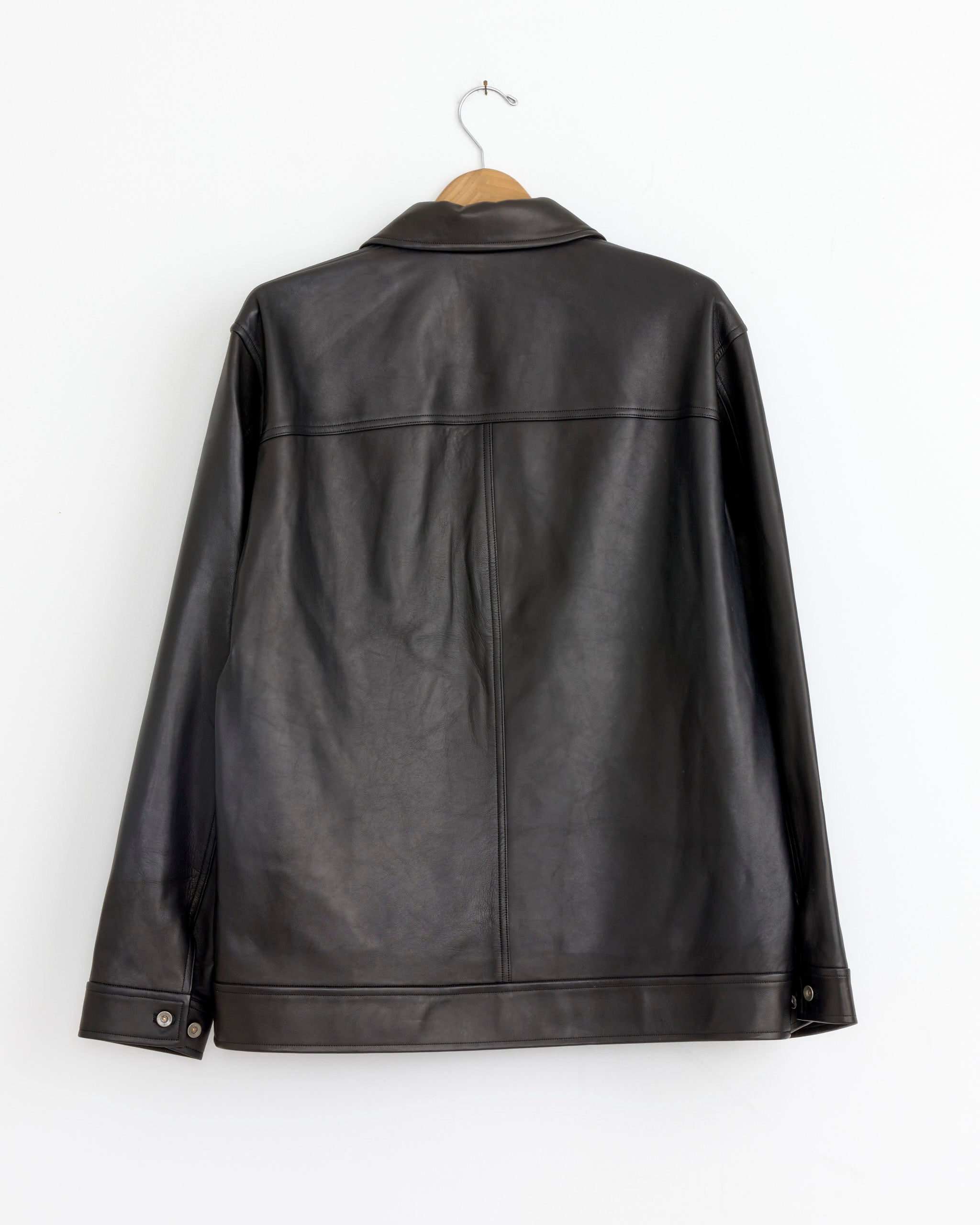 The Cale Glove Lamb Leather Jacket in Black, made in Japan, hangs on a wooden hanger against a white wall. Shown from the back, it features long sleeves, a collar, buttoned cuffs, and is available in size 4.