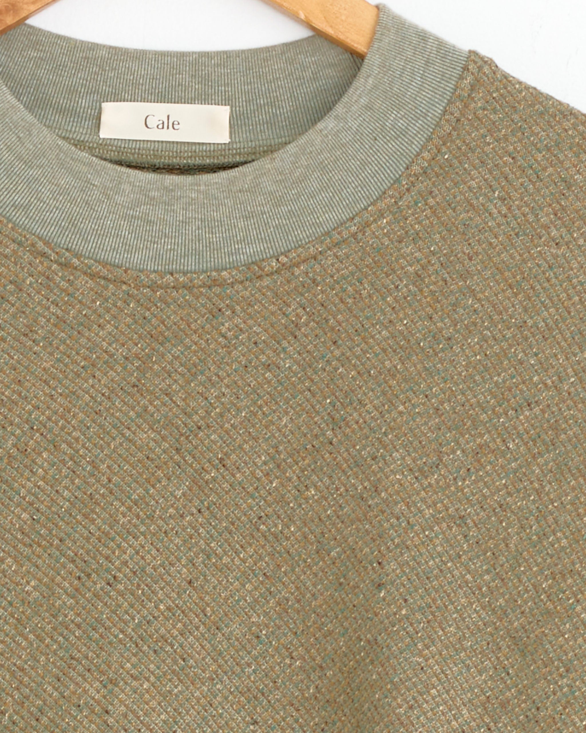 Close-up of the upper part of the Silk Cotton Jacquard Sweater in Beige by Cale on a wooden hanger, featuring a ribbed collar and label; classic knit fleece pullover made in Japan.