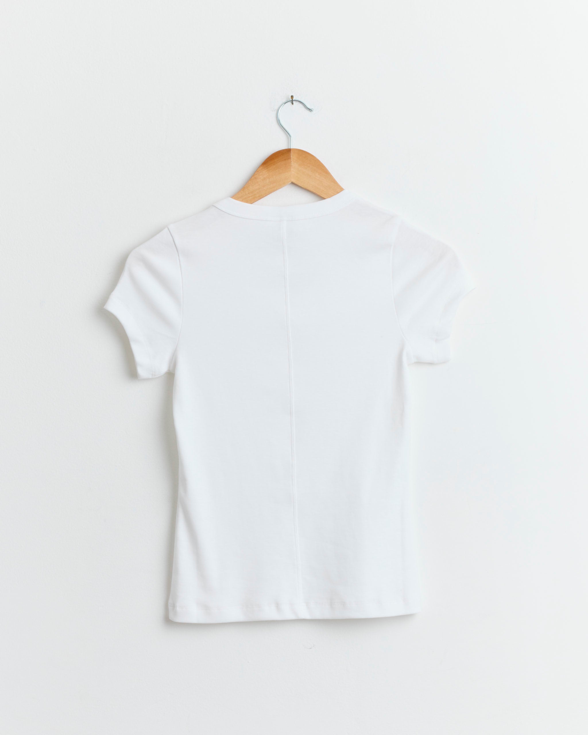 The FLORE FLORE Jill Baby Tee in White, made from organic cotton, is shown on a wooden hanger with its back facing a white wall.
