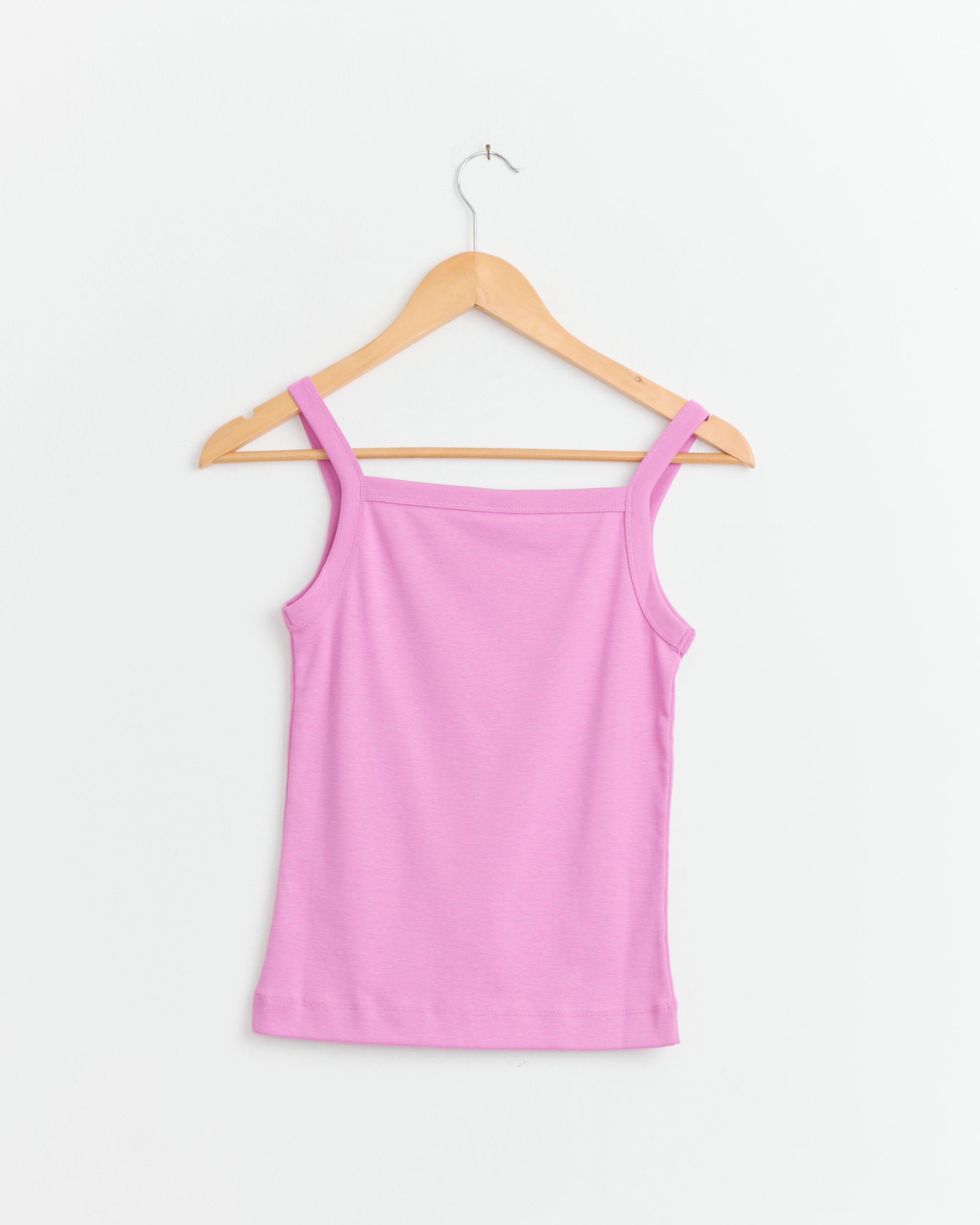 The FLORE FLORE May Cami in Crocus, featuring a light pink ribbed design and form-fitting silhouette, is displayed on a wooden hanger against a plain white background.