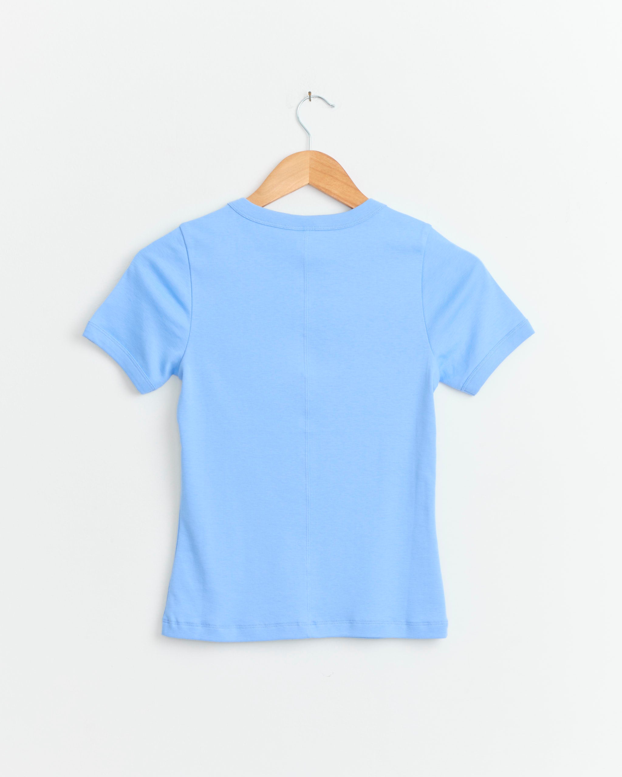 The FLORE FLORE Car Tee in Baby Blue, made from organic cotton with a fitted silhouette, is displayed on a wooden hanger against a white background, shown from the back.
