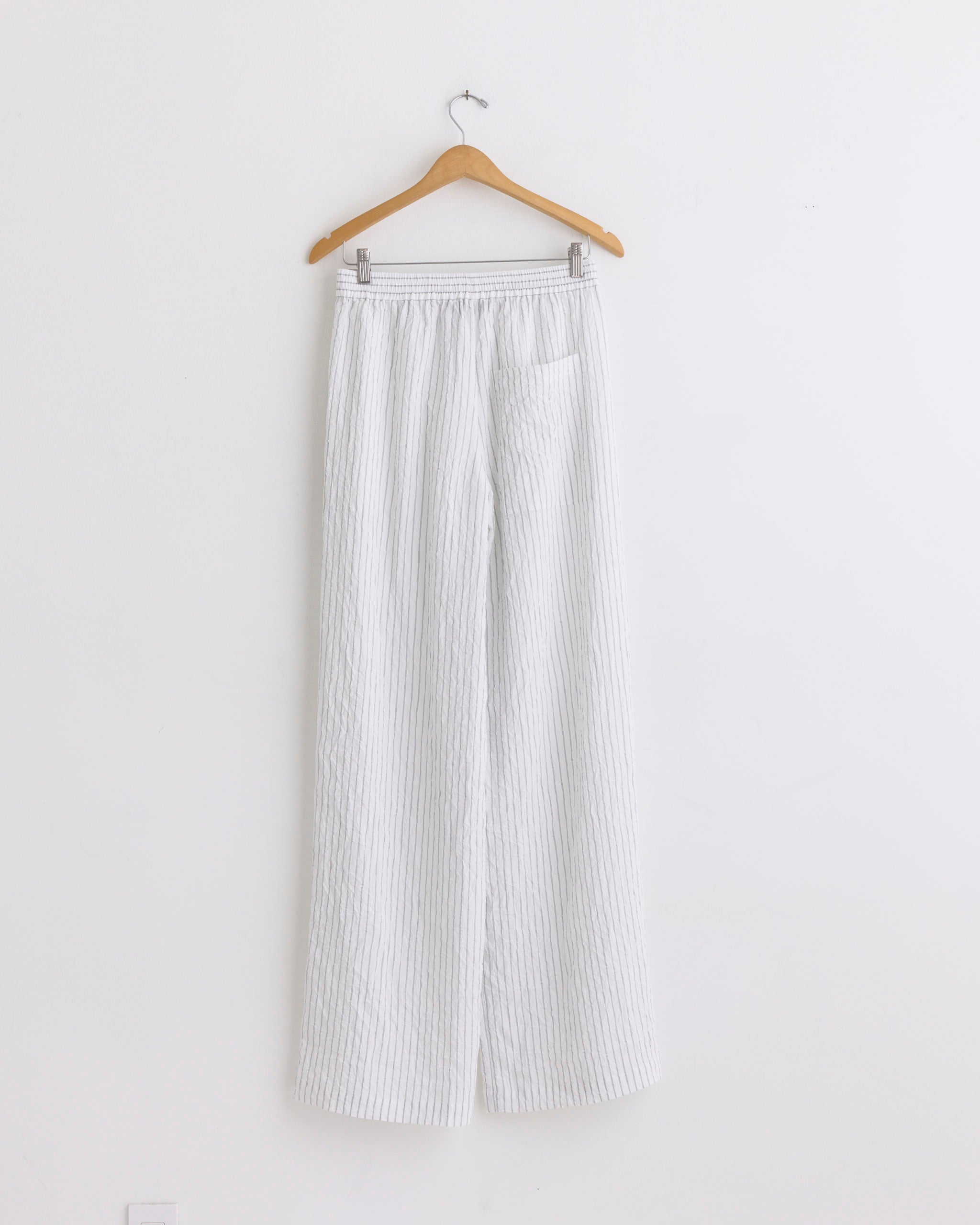 The Tibi Crinkle Stripe Wide Leg Pant in Ivory Multi, featuring a textured design and elastic waistband, is displayed on a wooden hanger against a plain white wall.