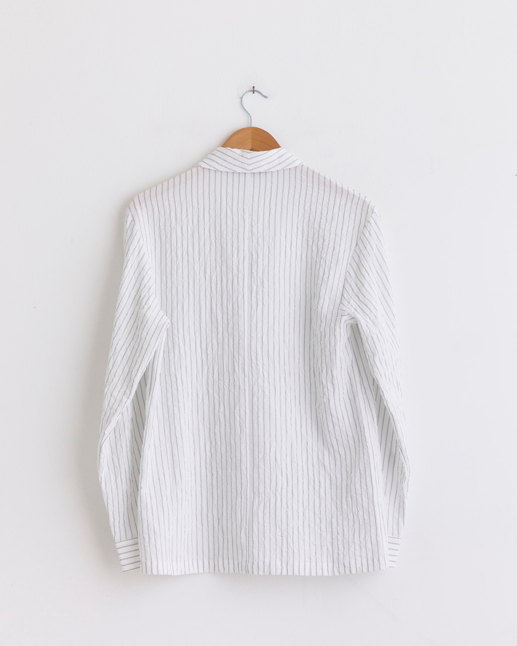 A Crinkle Stripe Shawl Collar Jacket in Ivory Multi by Tibi, shown from the back on a wooden hanger against a plain white wall—lightweight with thin vertical stripes and a women's blazer silhouette.