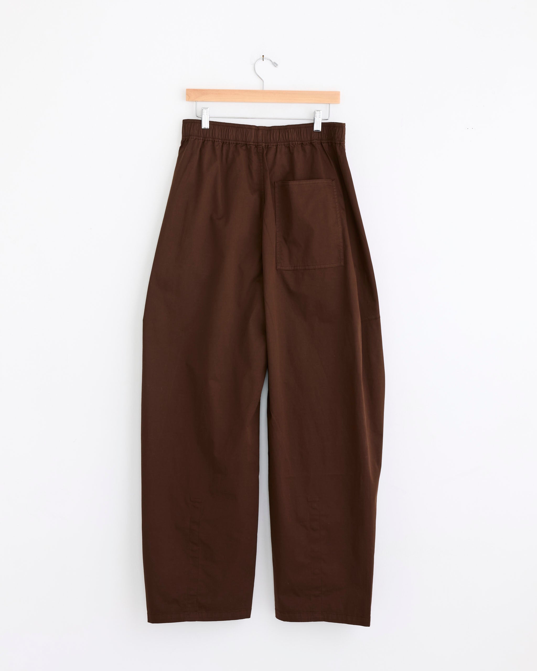 The Winslow Pant in Brown by Tibi features a loose, wide-leg cotton sateen fit, garment-dyed finish, elastic waistband, and a single back pocket, shown on a wooden hanger against a white background.