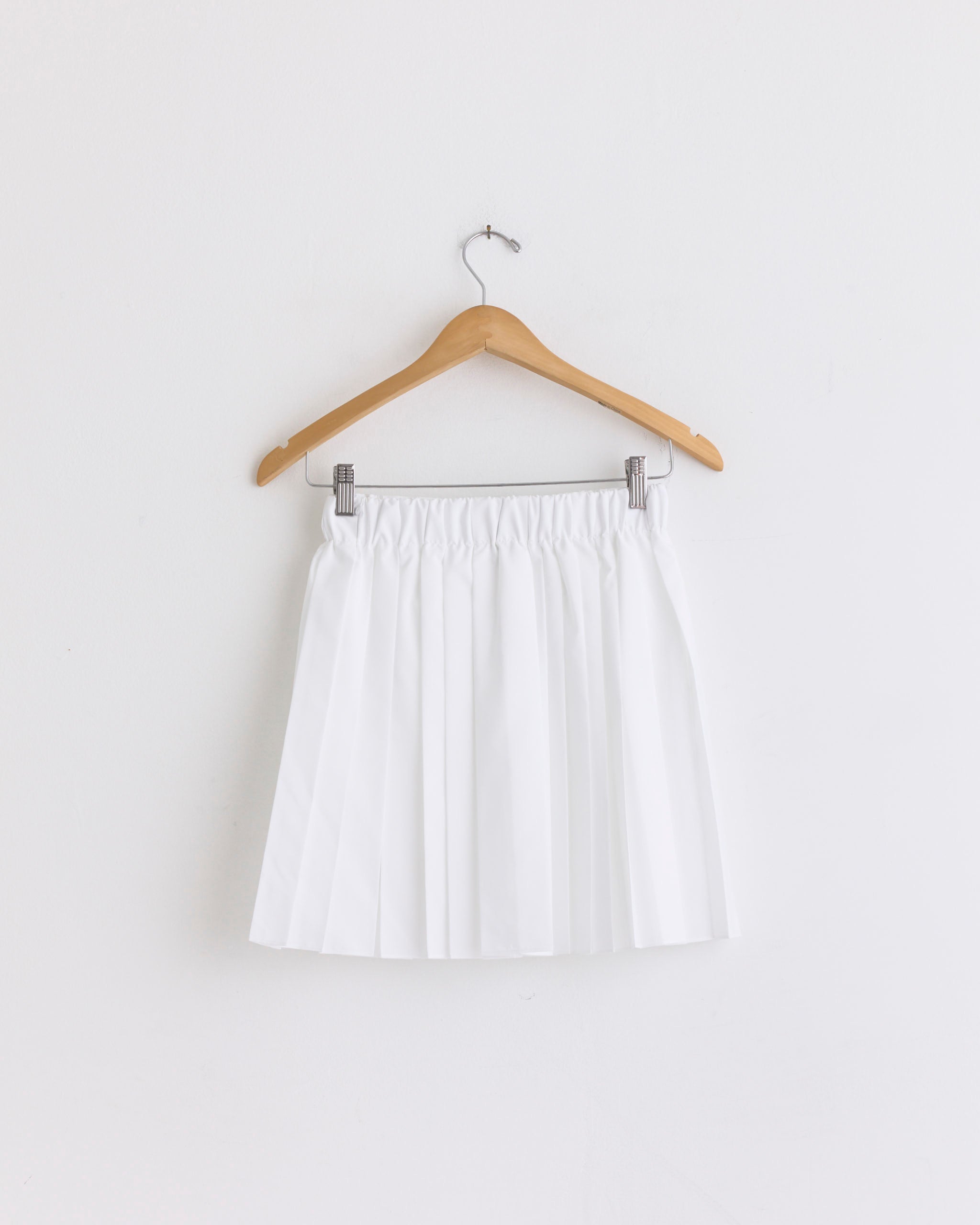 The Tibi Pull On Mini Skirt in White, featuring pleats and an elastic waistband, hangs on a wooden hanger against a plain white wall.