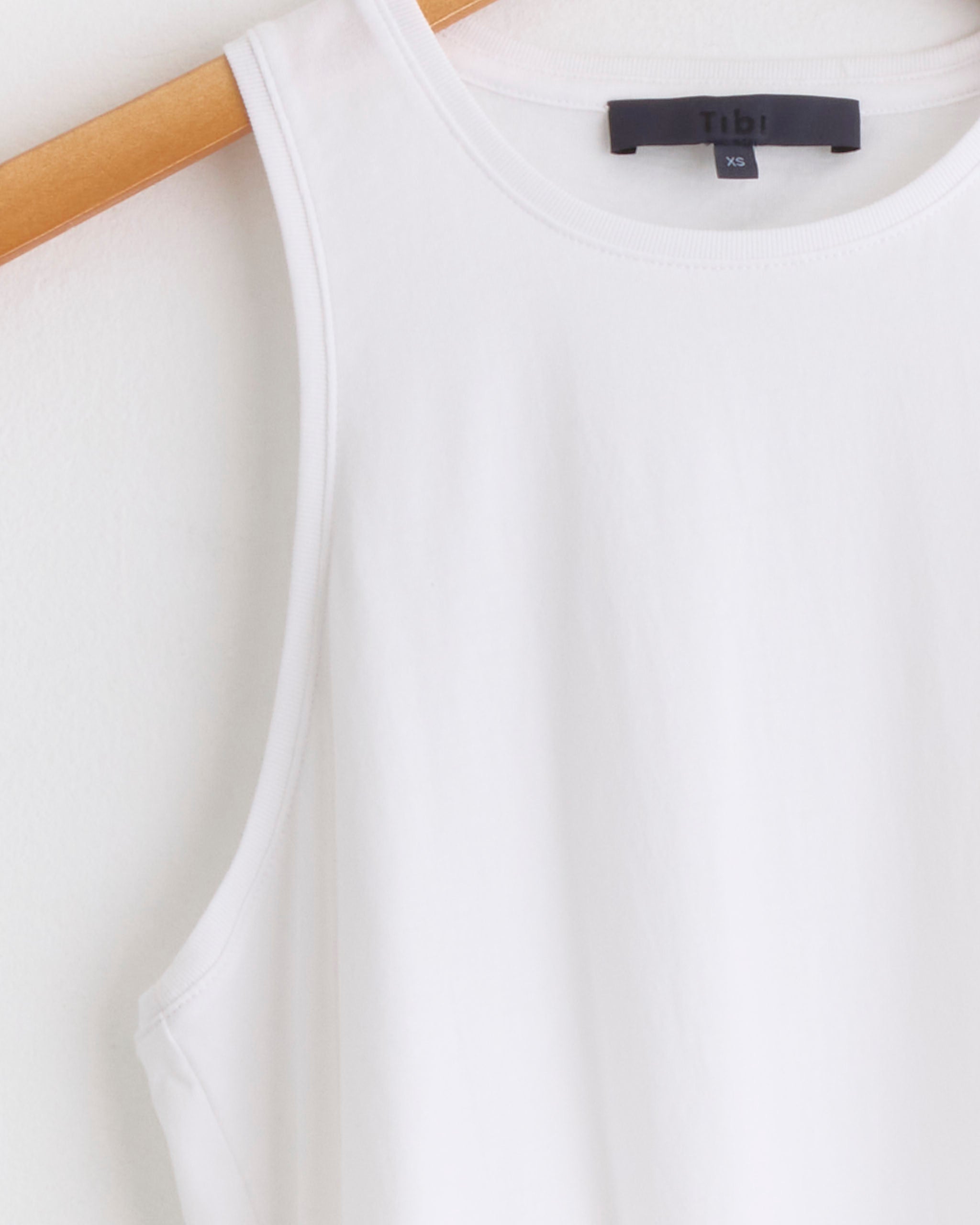 The Tibi Shirred Back Tank in White, size XS, hangs on a wooden hanger against a light-colored wall.
