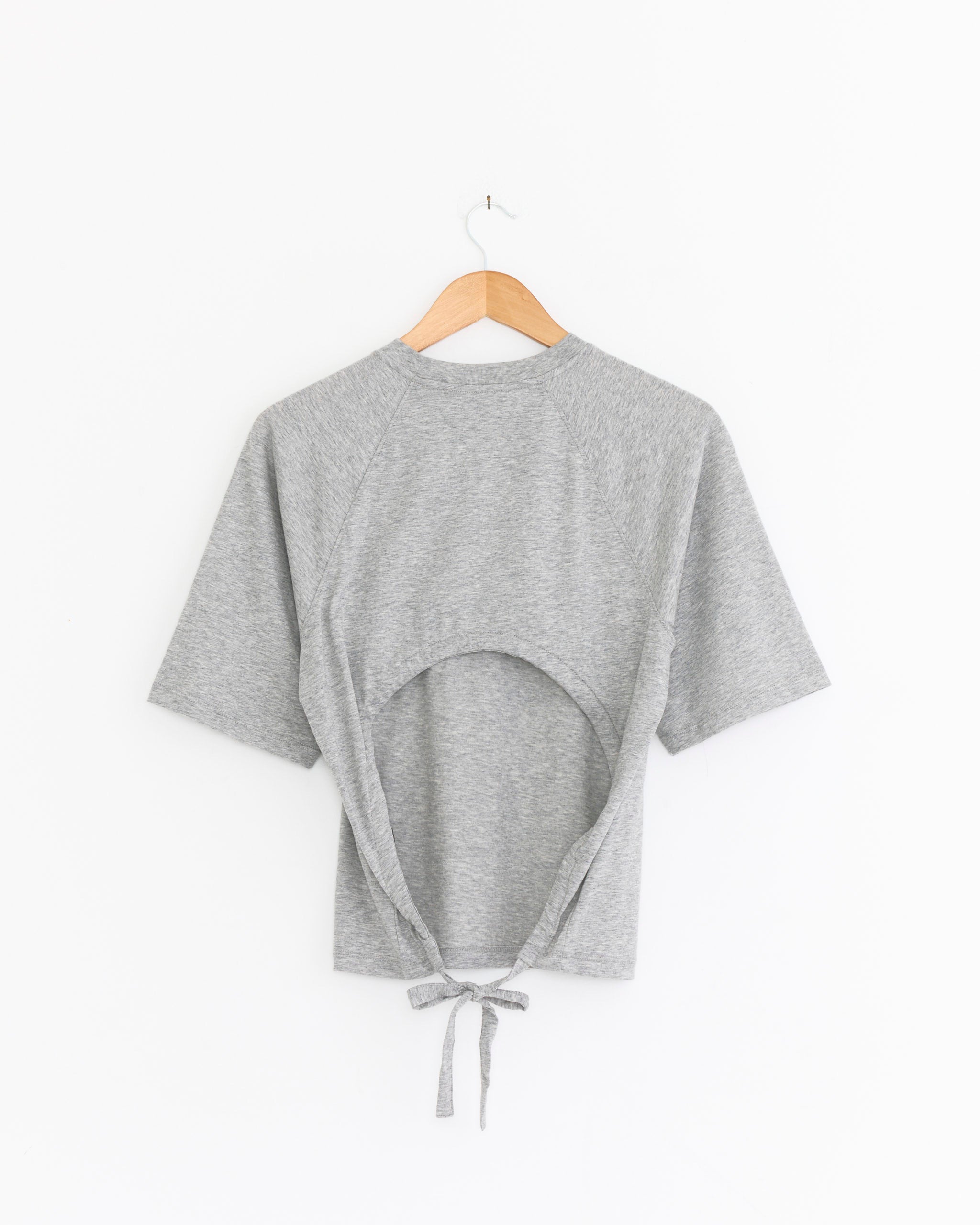 The Tibi Shirred Back Top in Heather Grey, shown on a wooden hanger against a white background, features short sleeves and a large circular back cutout with adjustable shirring and two ties at the bottom.