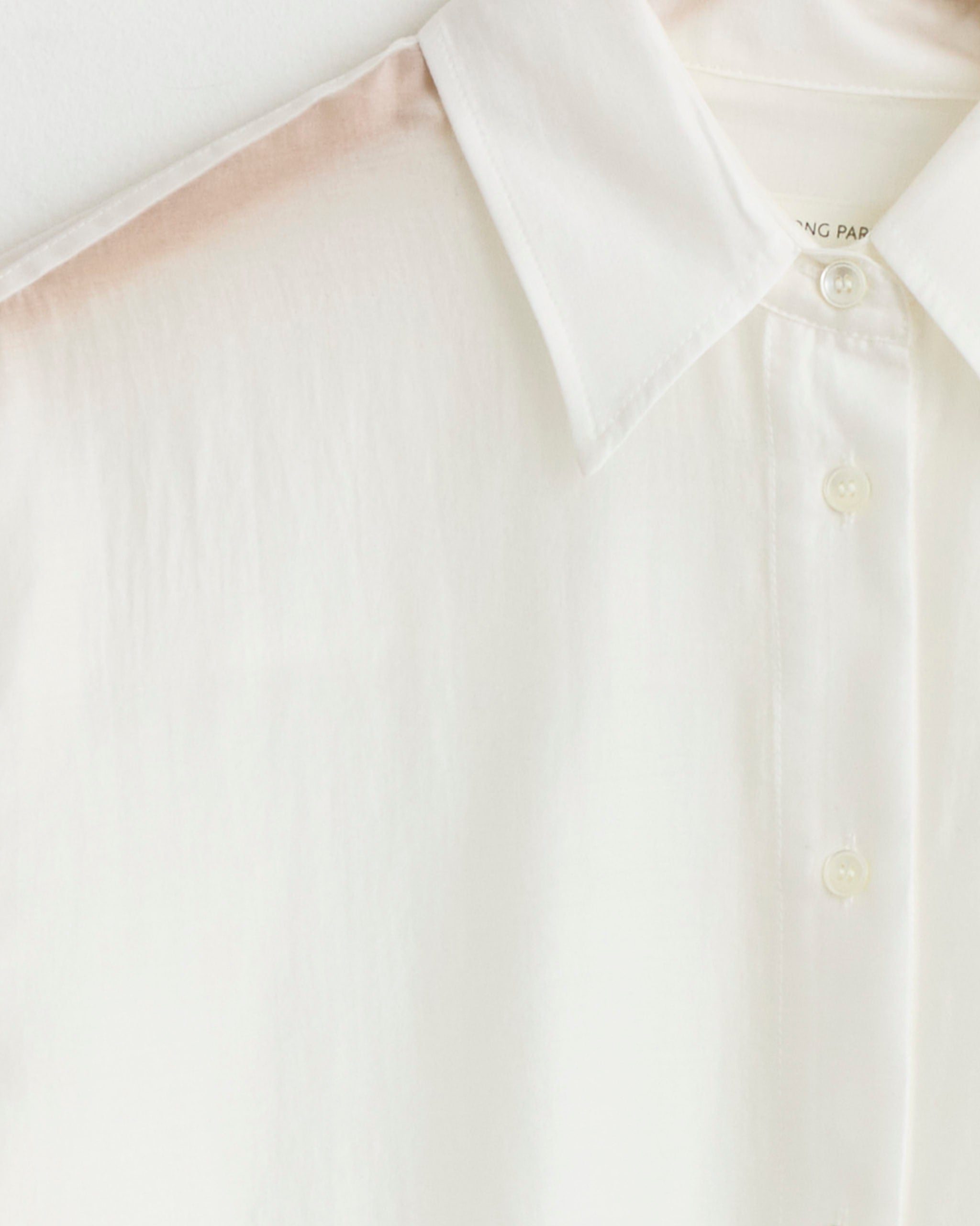 A close-up of the upper part of the Mijeong Park Semi Sheer Shirt in White, an oversized collared button-up made from a tencel blend, hanging on a hanger against a light background.