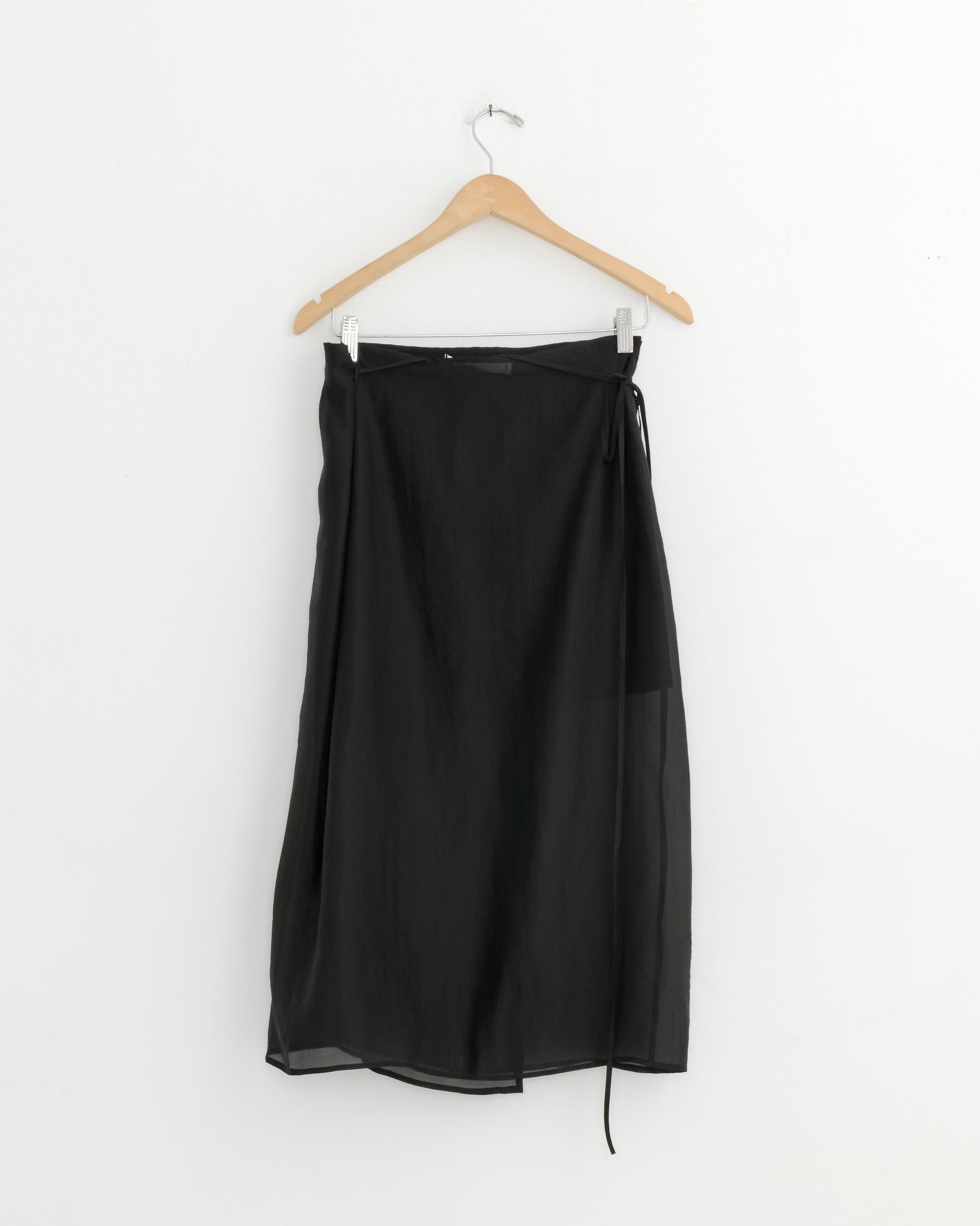 The Mijeong Park Organza Wrap Skirt in Black, a minimalist midi-length design with a side tie, is displayed on a wooden hanger against a plain white wall.