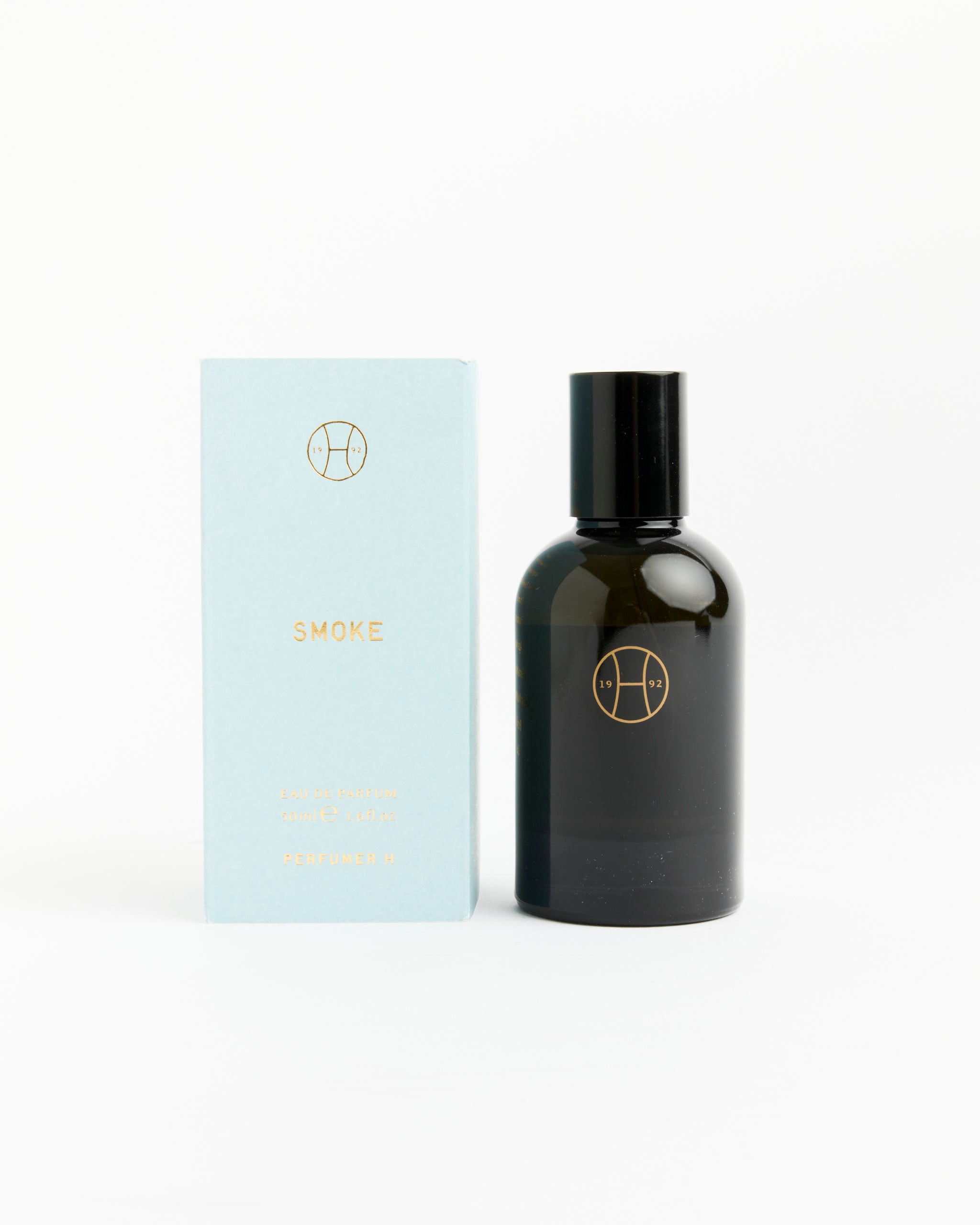 A light blue box labeled Smoke Perfume by Perfumer H sits beside a dark glass bottle with a black cap, both adorned with minimalist gold logos and white labels, evoking the amber and woody notes within.