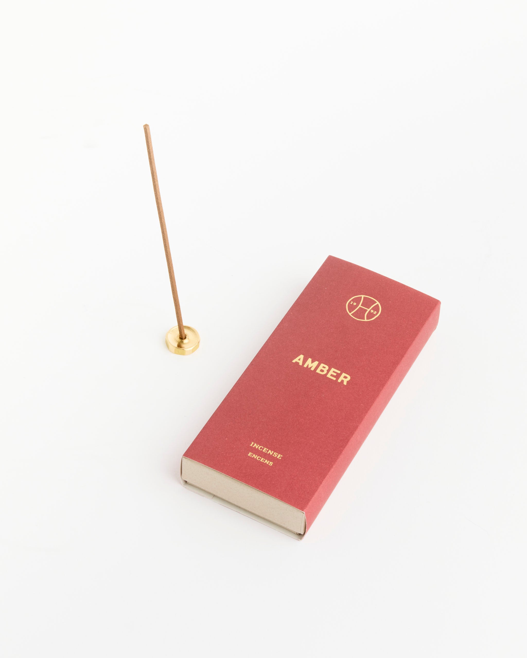 A stick of natural incense stands upright in a small, round brass holder beside a red Perfumer H Amber Incense box set against a white background.