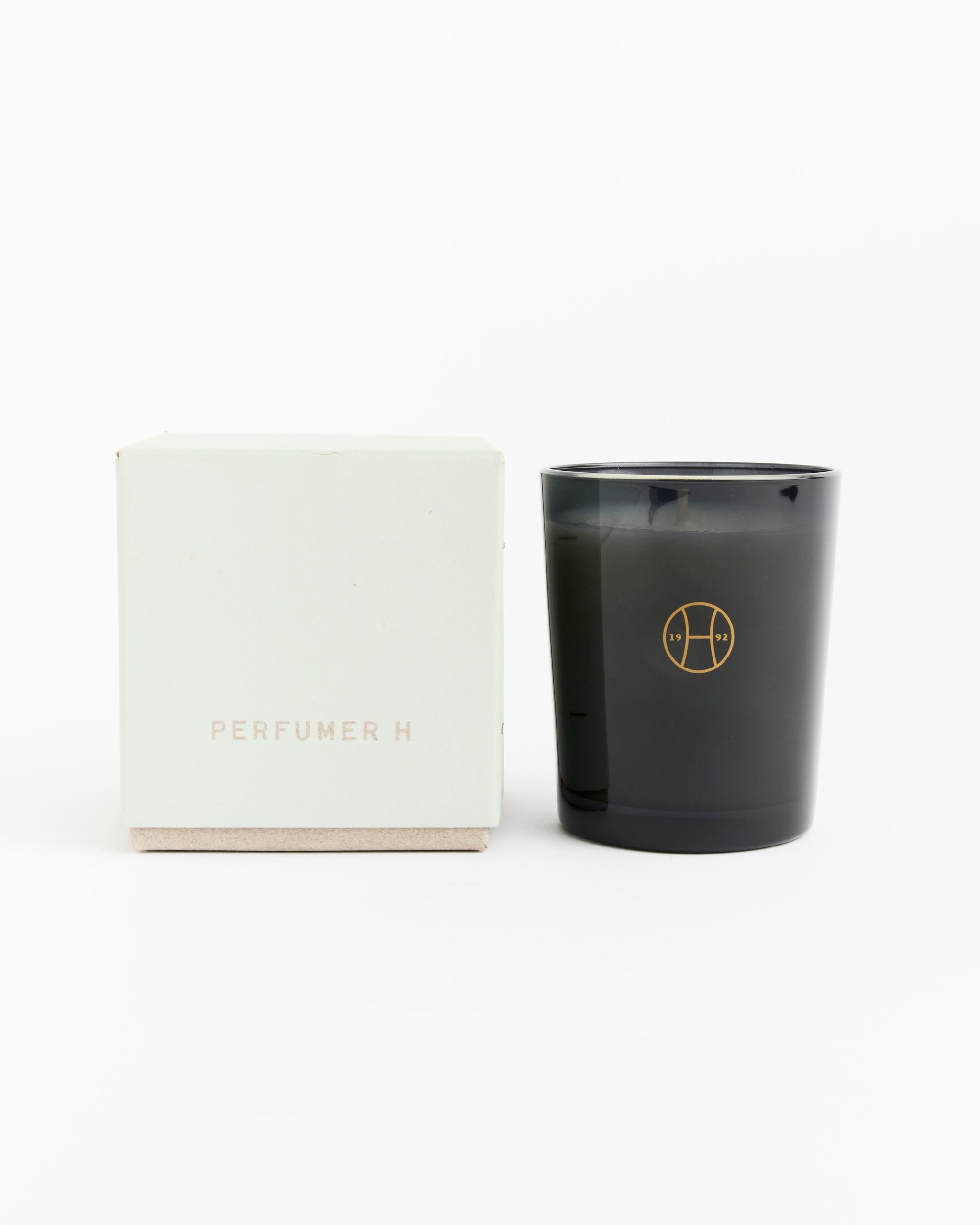 The Perfumer H Moss Candle, hand poured in a glass holder with a gold logo, rests beside its white PERFUMER H box on a plain white background.