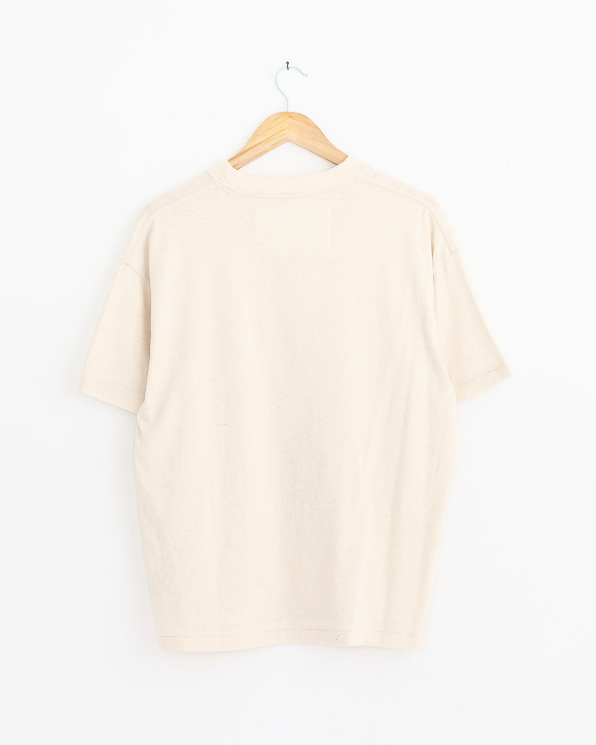 The Dana Lee Brown Boxy Tee in Undyed, a plain beige organic short-sleeve shirt, is displayed from the back on a wooden hanger against a white background.