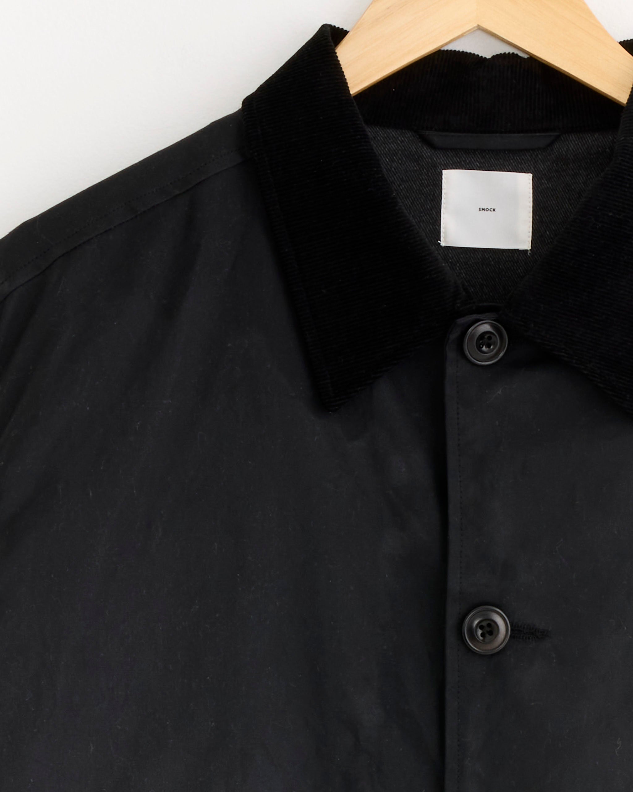 Close-up of the SMOCK Blakey Jacket in Waxed Cotton Black, featuring a black corduroy collar, displayed on a wooden hanger against a white background.