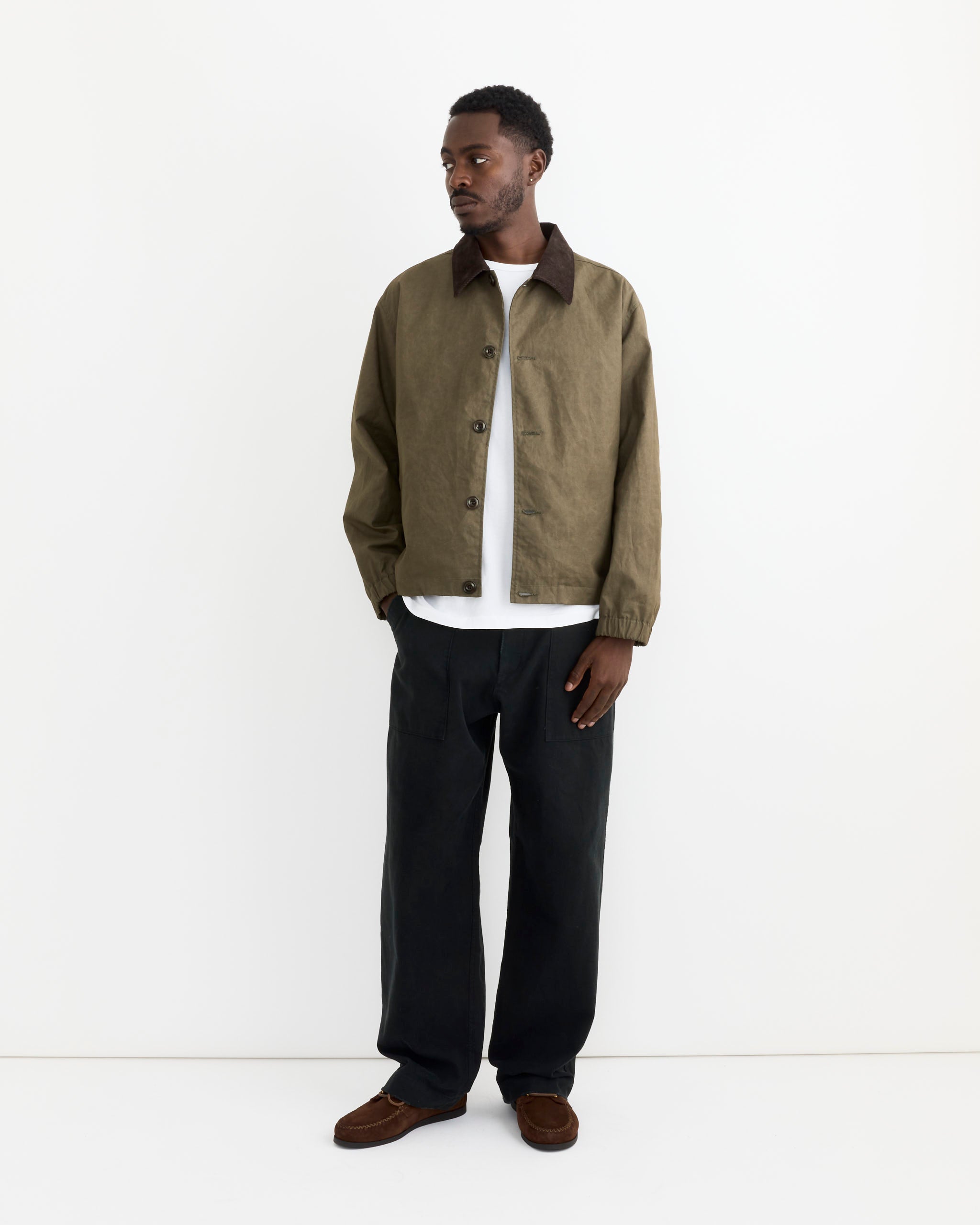 A man stands against a white background wearing an olive green jacket, brown shoes, and SMOCK Toyon Pant in French Cotton Twill Black. He gazes slightly left, embodying an effortlessly utilitarian, workwear-inspired style.
